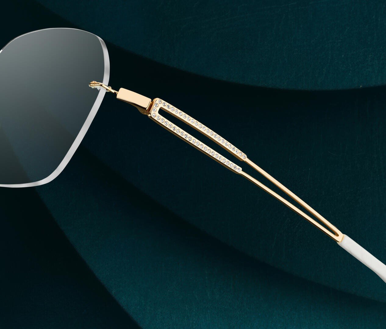 Nahaufnahme einer Brille mit gold- und diamantbesetztem Bügel vor einem dunkelgrünen, strukturierten Hintergrund. Das Bild hebt die luxuriösen Details und die Handwerkskunst des Brillenbügels hervor.