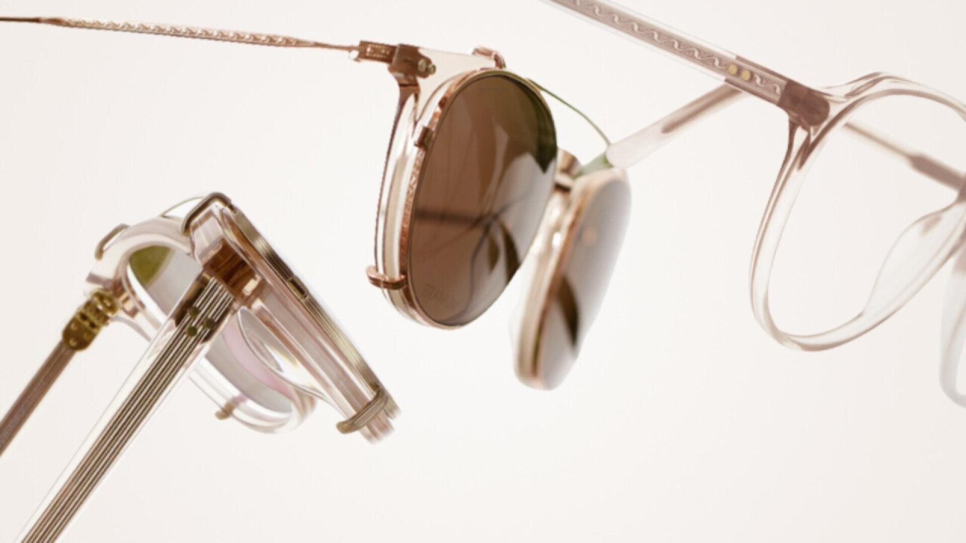 Close-up of two pairs of eyeglasses, one with attached brown clip-on sunglasses lenses, both with transparent frames, set against a light, neutral background.