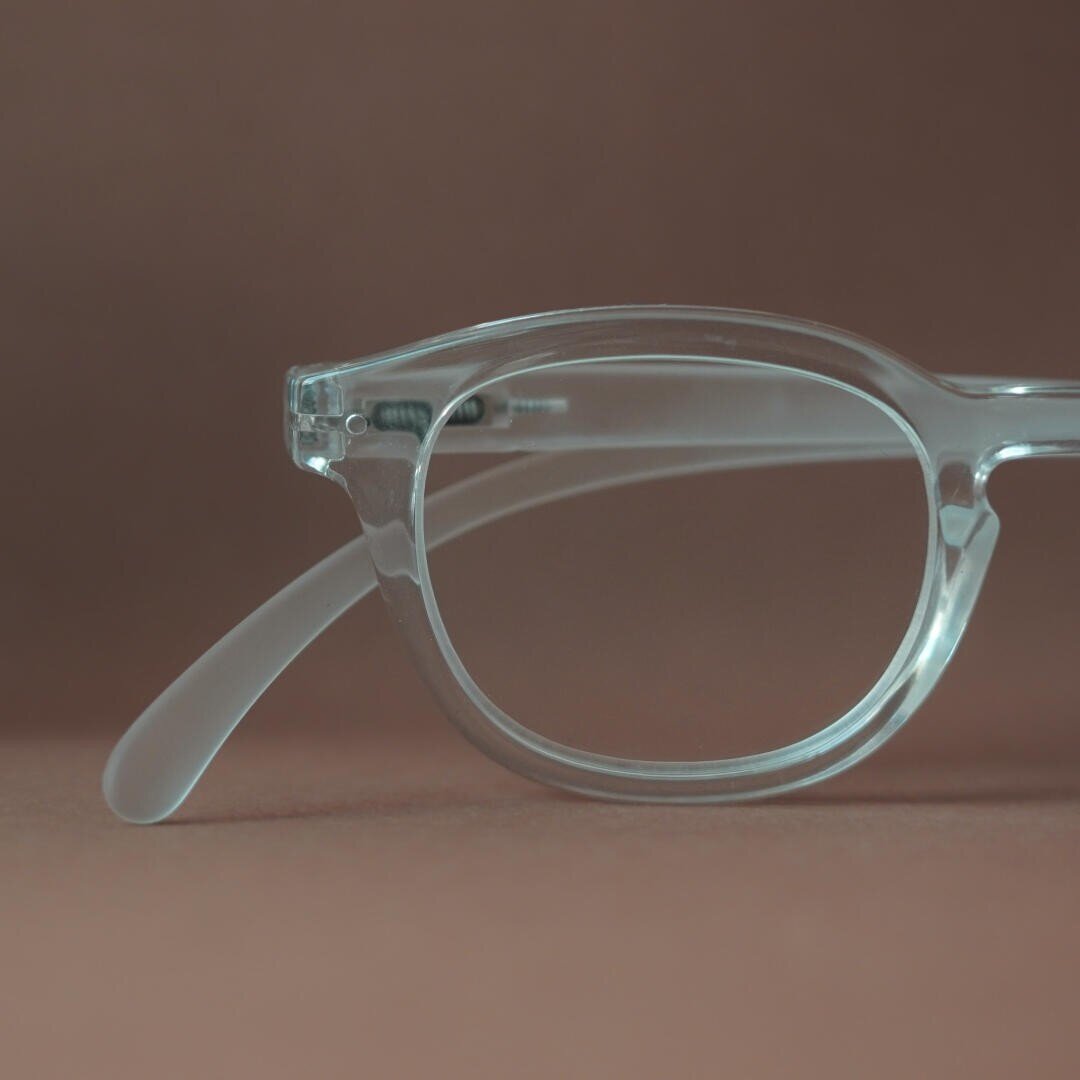 Nahaufnahme eines transparenten Brillengestells mit klaren Gläsern und Bügeln vor einem schlichten, braunen Hintergrund. Nur die linke Hälfte der Brille ist sichtbar.