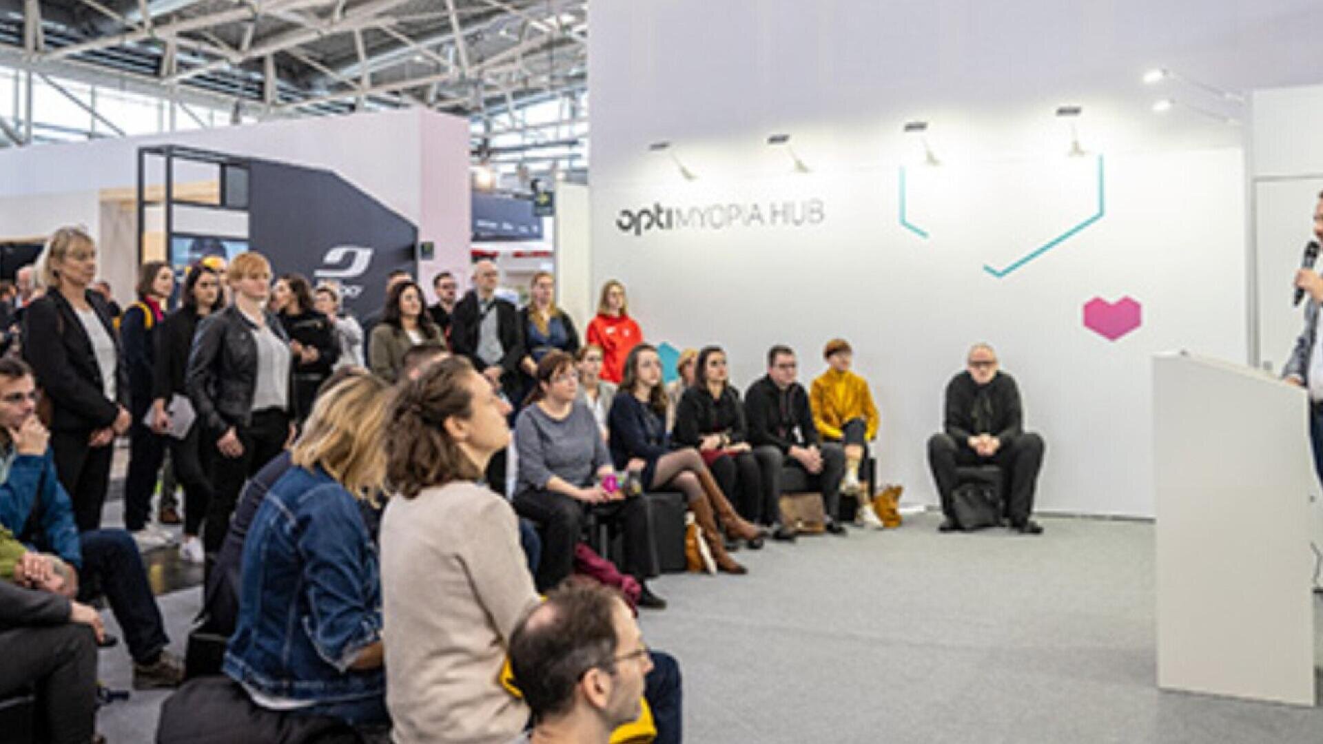 A group of people sits and stands attentively in a modern indoor space, listening to a speaker at an event. The area has white walls, bright lighting, and signage reading optimum HUB.
