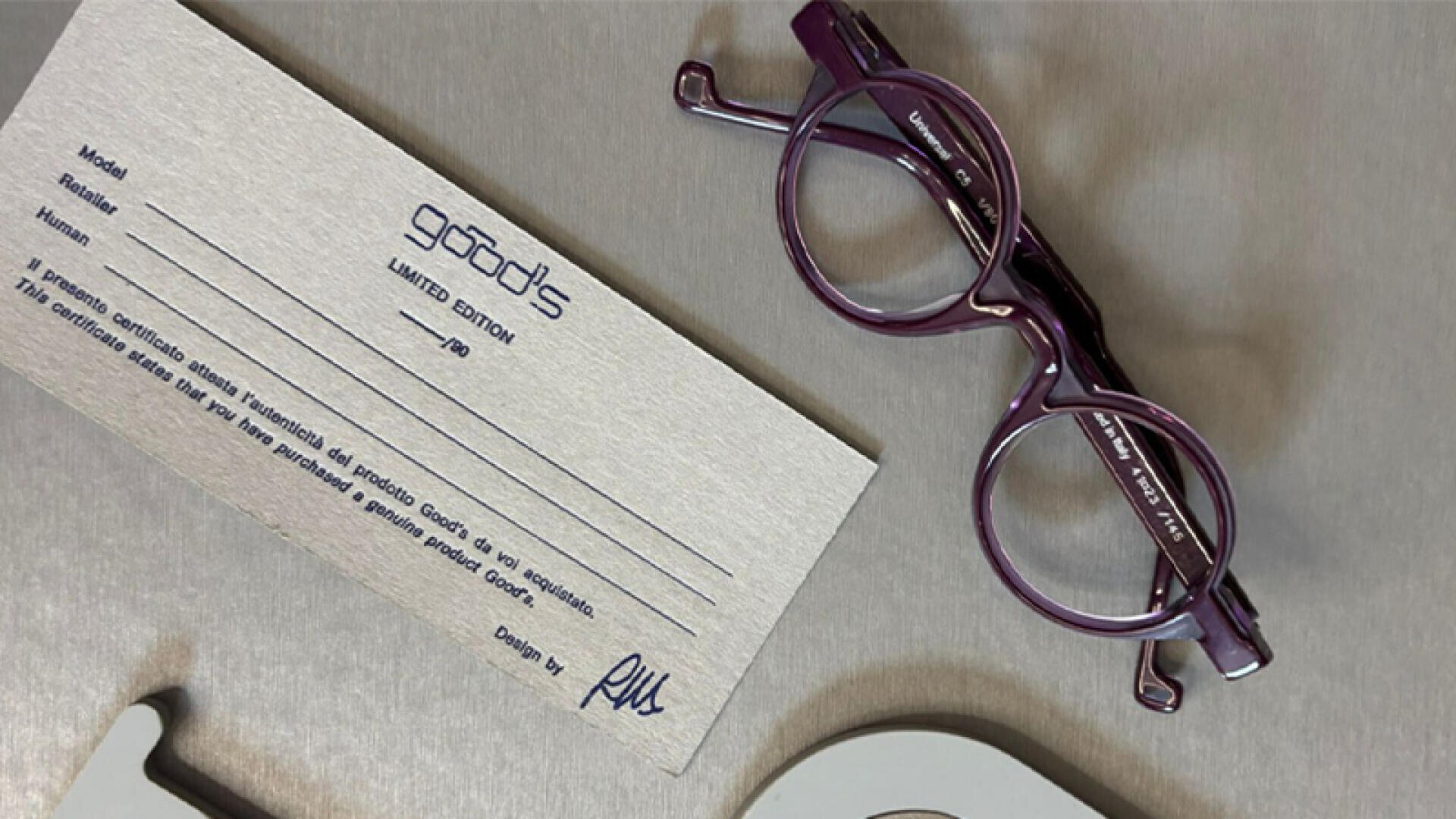 A pair of round, purple glasses lies next to a limited edition certificate card on a gray surface. The certificate contains text and a signature, partly in Italian.