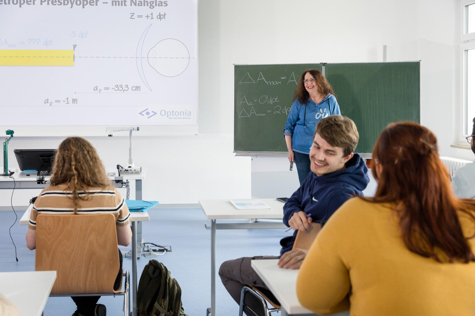 Eine Lehrerin steht an einer Tafel und lächelt den Schülern zu, die in einem hellen Klassenzimmer an ihren Tischen sitzen. Auf eine Leinwand hinter ihr werden physikalische Diagramme projiziert.