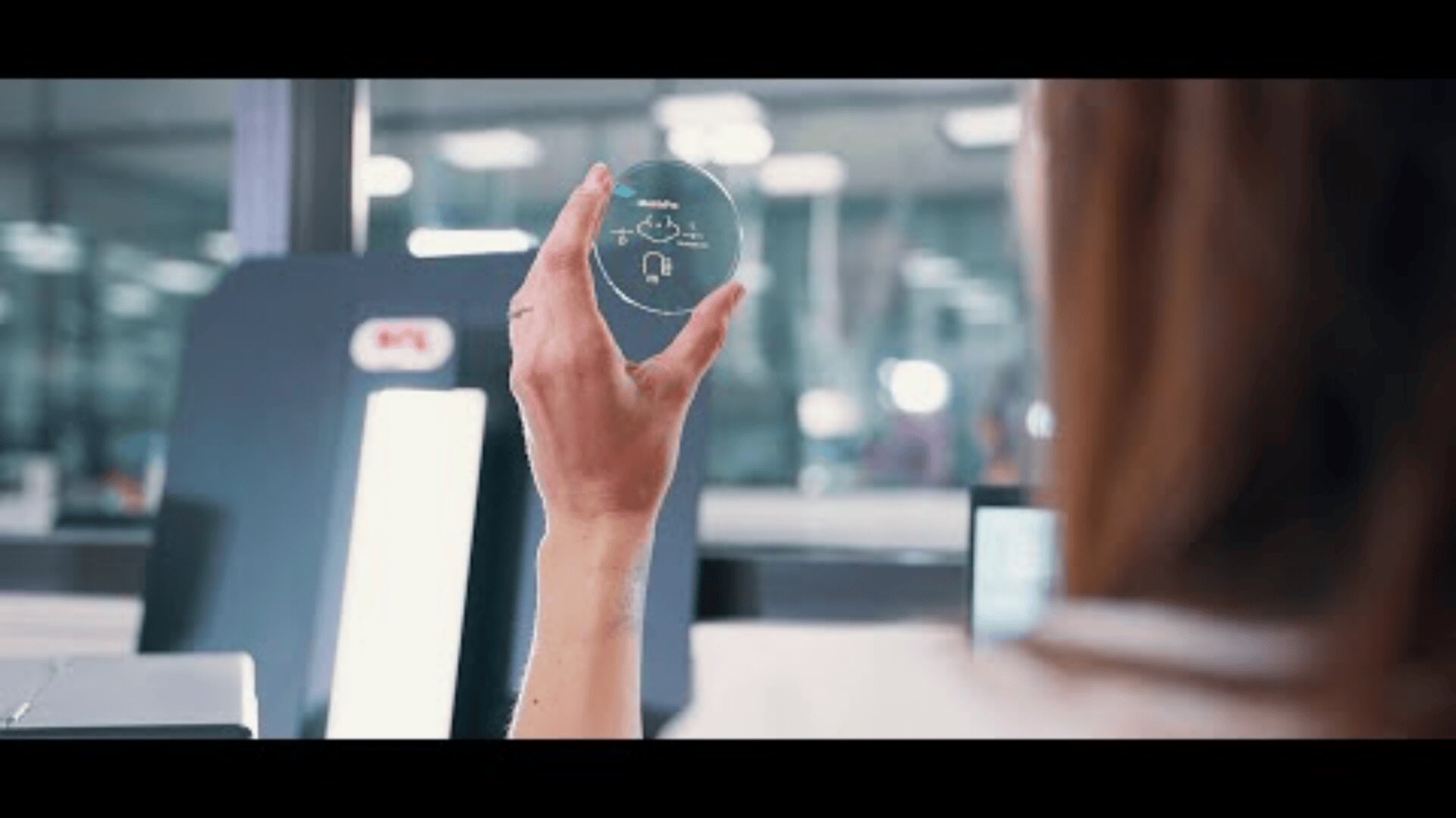 In einer hellen, modernen Labor- oder Produktionsumgebung hält eine Person eine transparente, runde Linse oder ein Glas mit kleinen, weißen, aufgedruckten technischen Symbolen hoch.