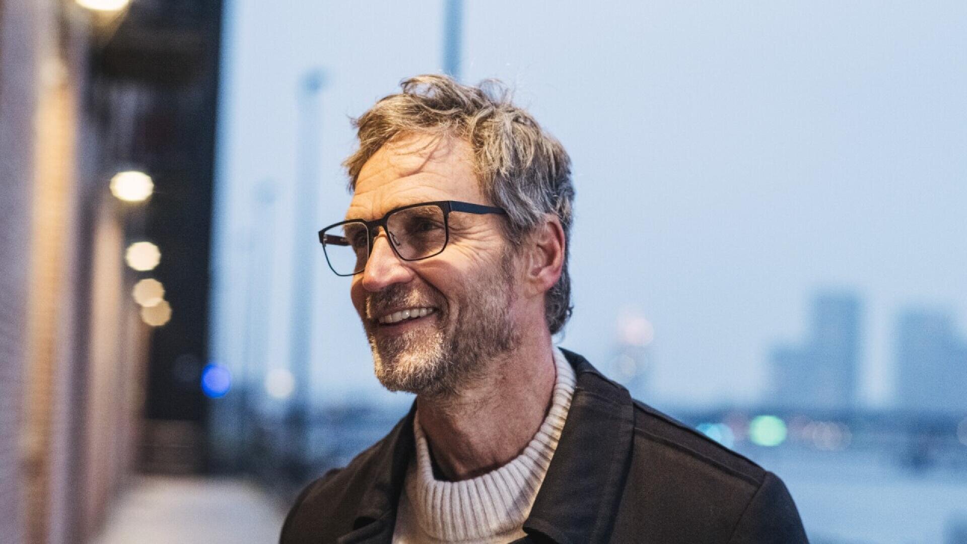 A middle-aged man with glasses and gray hair smiles while standing outdoors in the evening, dressed in a sweater and jacket, with blurred city lights and buildings in the background.