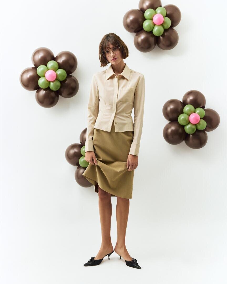 A woman in a beige blouse and khaki skirt stands in front of a white wall decorated with four flower-shaped balloon arrangements in brown, green and pink. She is wearing glasses and black heels.