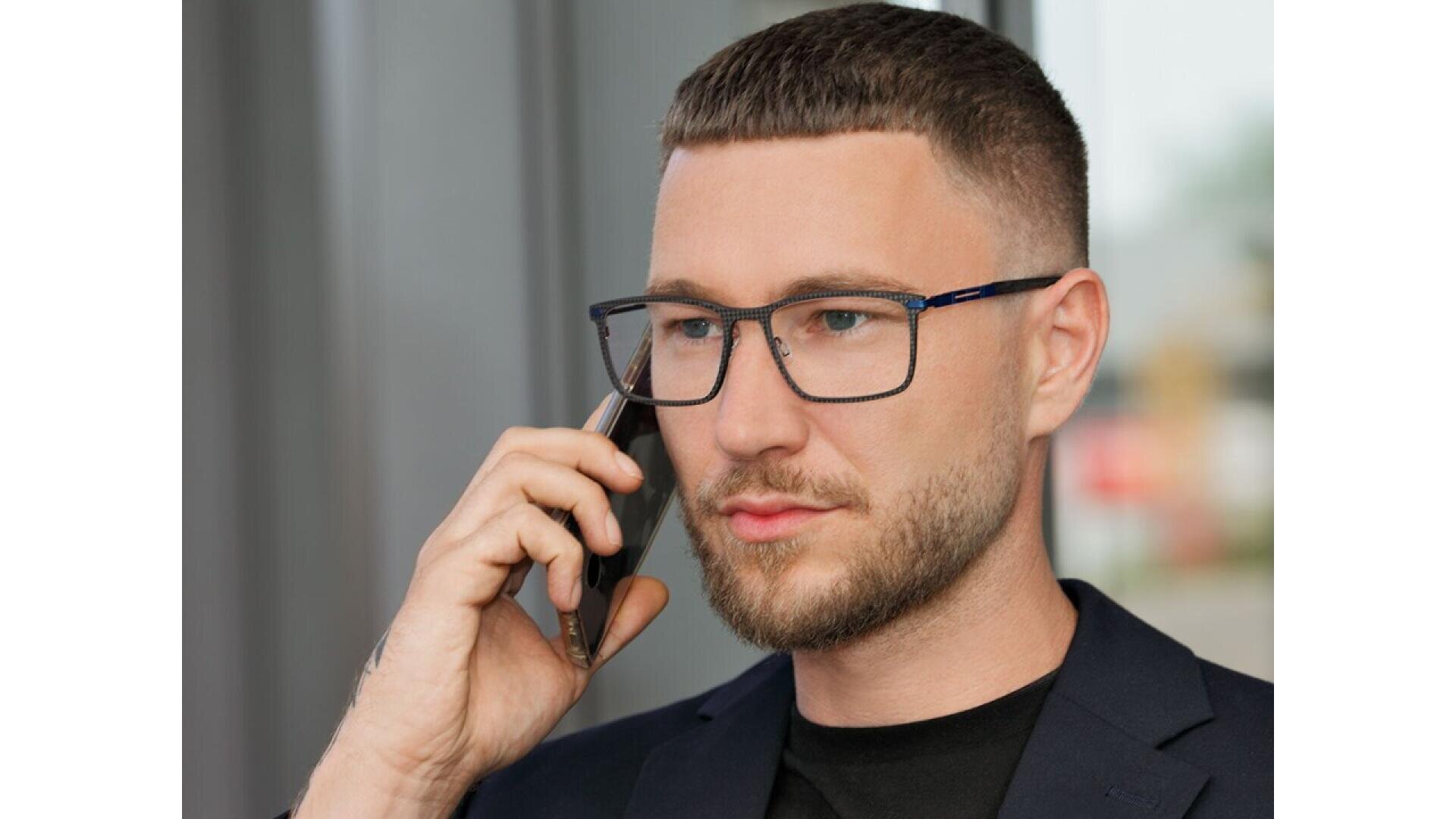 A man with short brown hair, a beard, and glasses is wearing a dark blazer and black shirt while talking on a smartphone, looking thoughtful.
