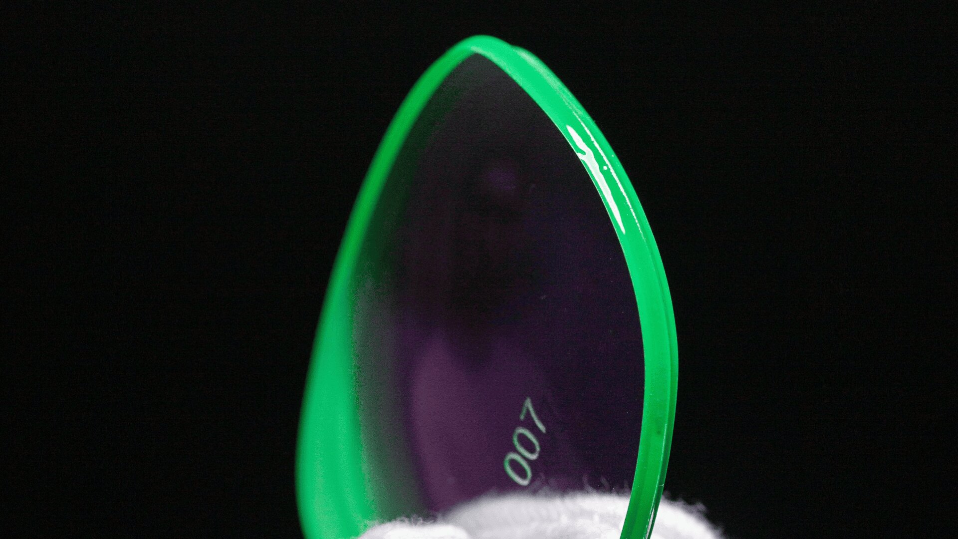 Nahaufnahme einer aufrecht gehaltenen, grün umrandeten, halbtransparenten Linse vor schwarzem Hintergrund, auf deren Oberfläche die Nummer 007 und ein kleines weißes Logo zu sehen sind.