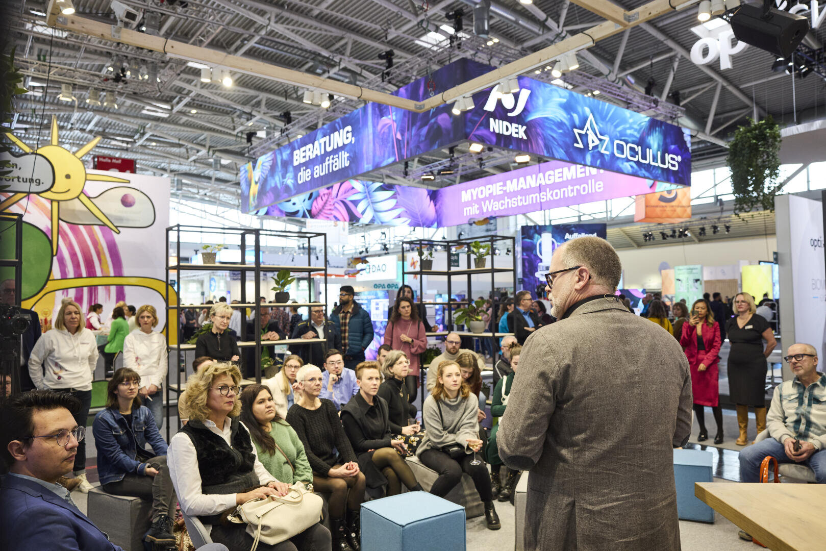Eine Gruppe von Menschen sitzt auf einer Messe oder Konferenz da und hört einem Redner aufmerksam zu. Im Hintergrund sind bunte Banner und Stände zu sehen und über ihnen herrscht eine moderne Beleuchtung.