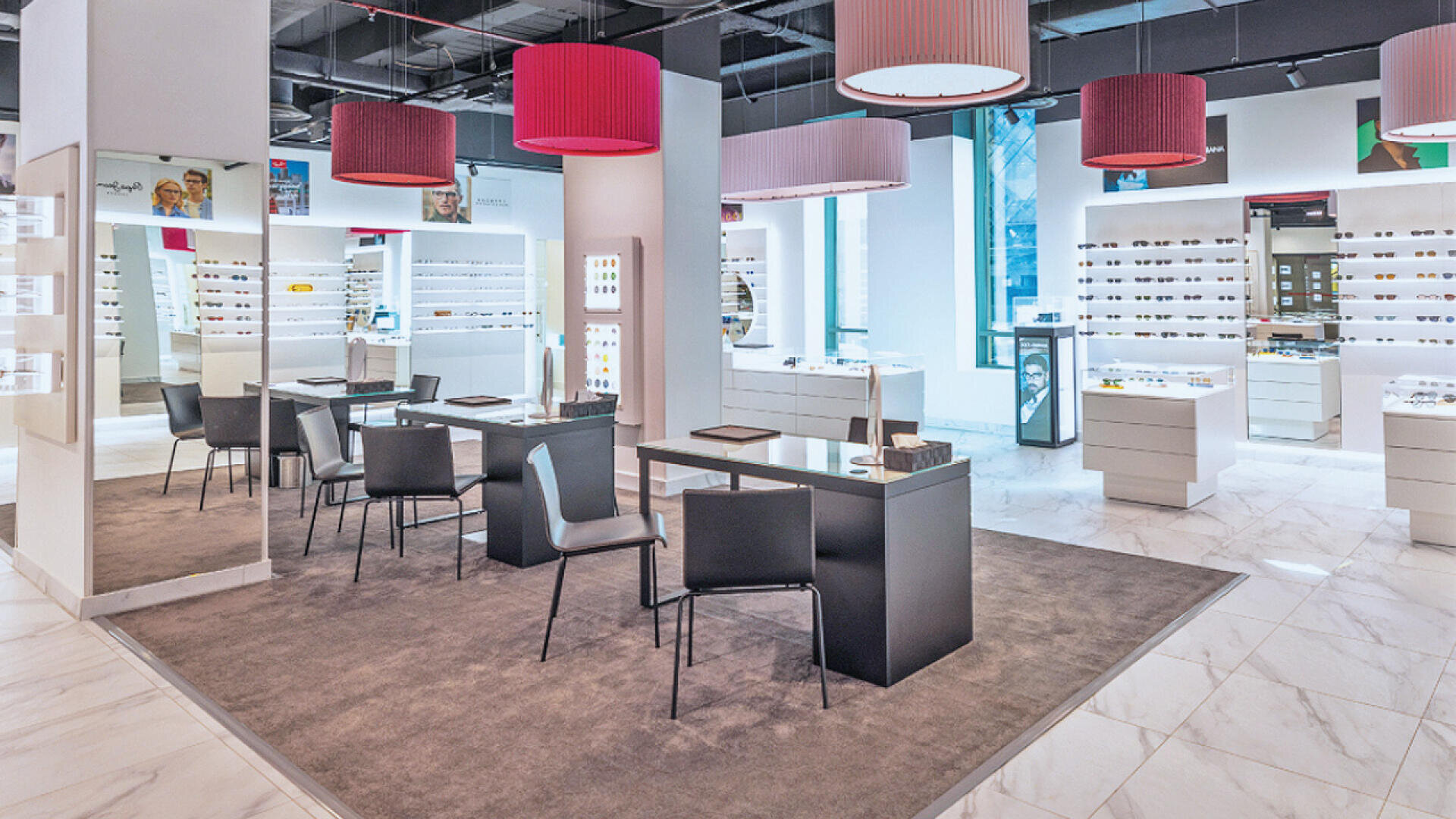 A modern, brightly lit eyewear store with display shelves of glasses, sleek tables and chairs on a gray rug, large ceiling lights with pink shades, and white tiled floors.
