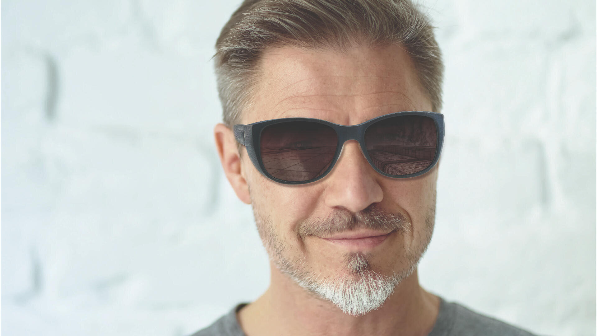 Ein Mann mittleren Alters mit kurzem, hellbraunem Haar und ordentlich gestutztem Bart, einer dunklen Sonnenbrille und einem grauen Hemd, der vor einem hellen, strukturierten Hintergrund leicht lächelt.