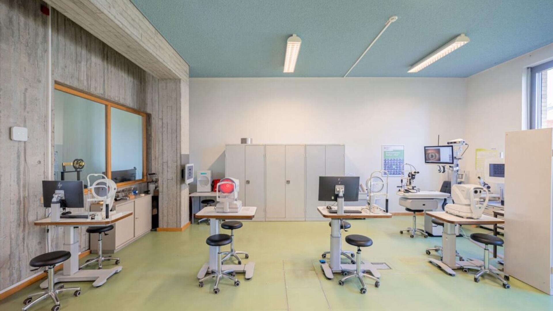 A bright, modern eye examination room with multiple vision testing machines, stools, computers, and medical equipment arranged neatly on light green flooring under fluorescent ceiling lights.