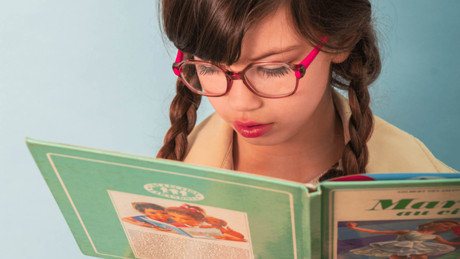 A young girl with braided hair and red glasses is reading a green book with an illustration and text on its cover, against a light blue background.