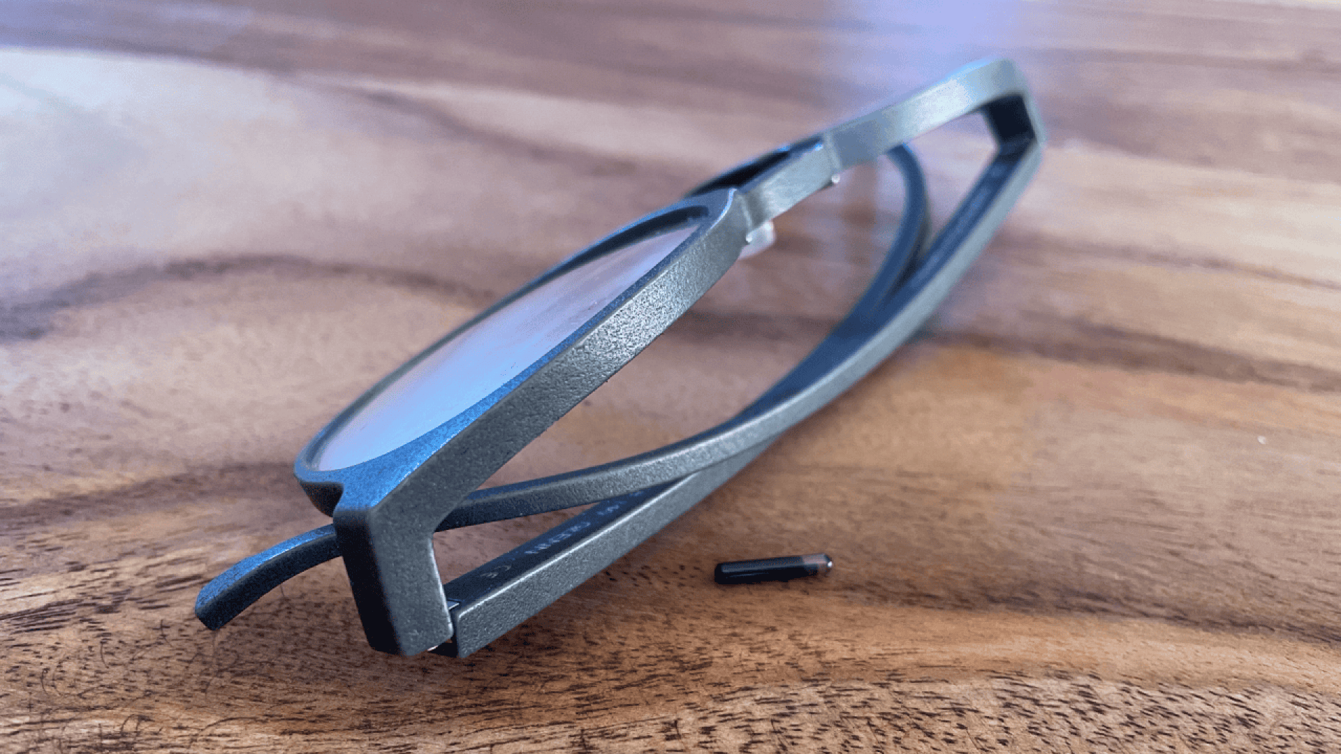 A pair of black eyeglasses with one of the arms detached, lying on a wooden surface. A small screw, likely from the hinge, is positioned near the frame.