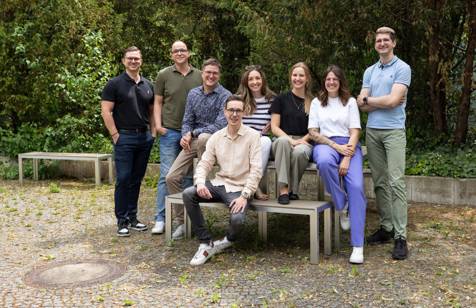 Acht Personen posieren und lächeln im Freien in legerer Kleidung; fünf stehen dahinter und drei sitzen auf Bänken. Bäume und Grünpflanzen bilden einen natürlichen Hintergrund. Die Atmosphäre wirkt freundlich und entspannt.