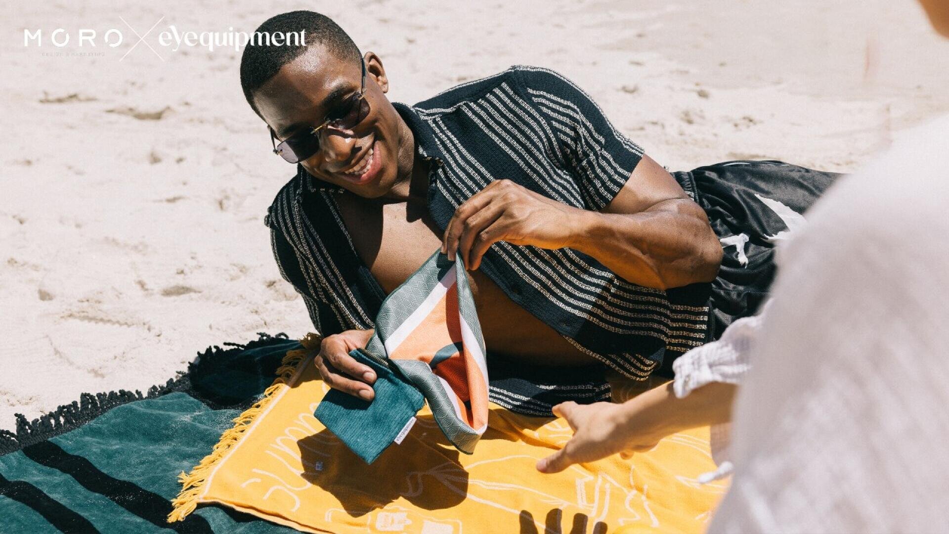 A man in sunglasses smiles while lying on a beach towel, holding colorful fabric. He is on a sandy beach with another person’s hand reaching toward him. The sun is shining brightly, creating a relaxed atmosphere.