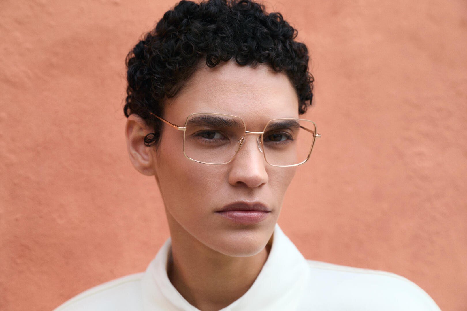 A person with short, curly dark hair and gold wire-frame glasses looks serious, standing against a peach-colored textured wall. They are wearing a white collared shirt.