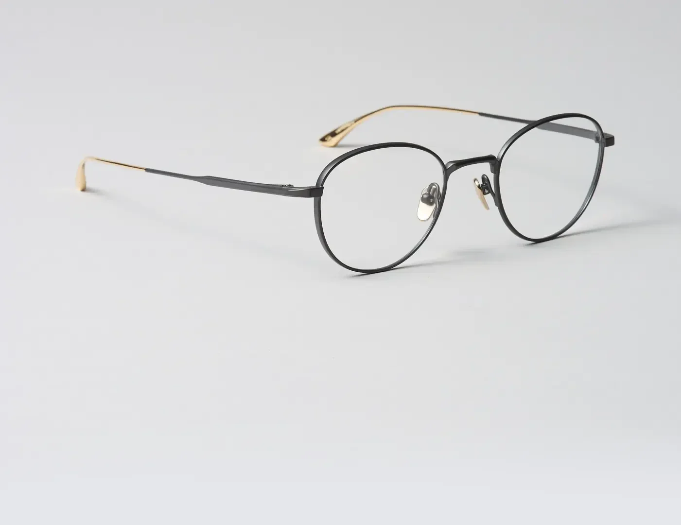 A pair of eyeglasses with thin black round frames and gold temples, resting on a plain light gray surface.