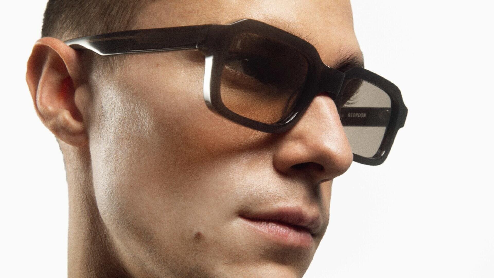 Eine Nahaufnahme einer Person mit einer großen, geometrischen dunklen Sonnenbrille, die leicht zur Seite blickt. Der Hintergrund ist schlicht weiß, die Person hat kurze Haare und glatte Haut.