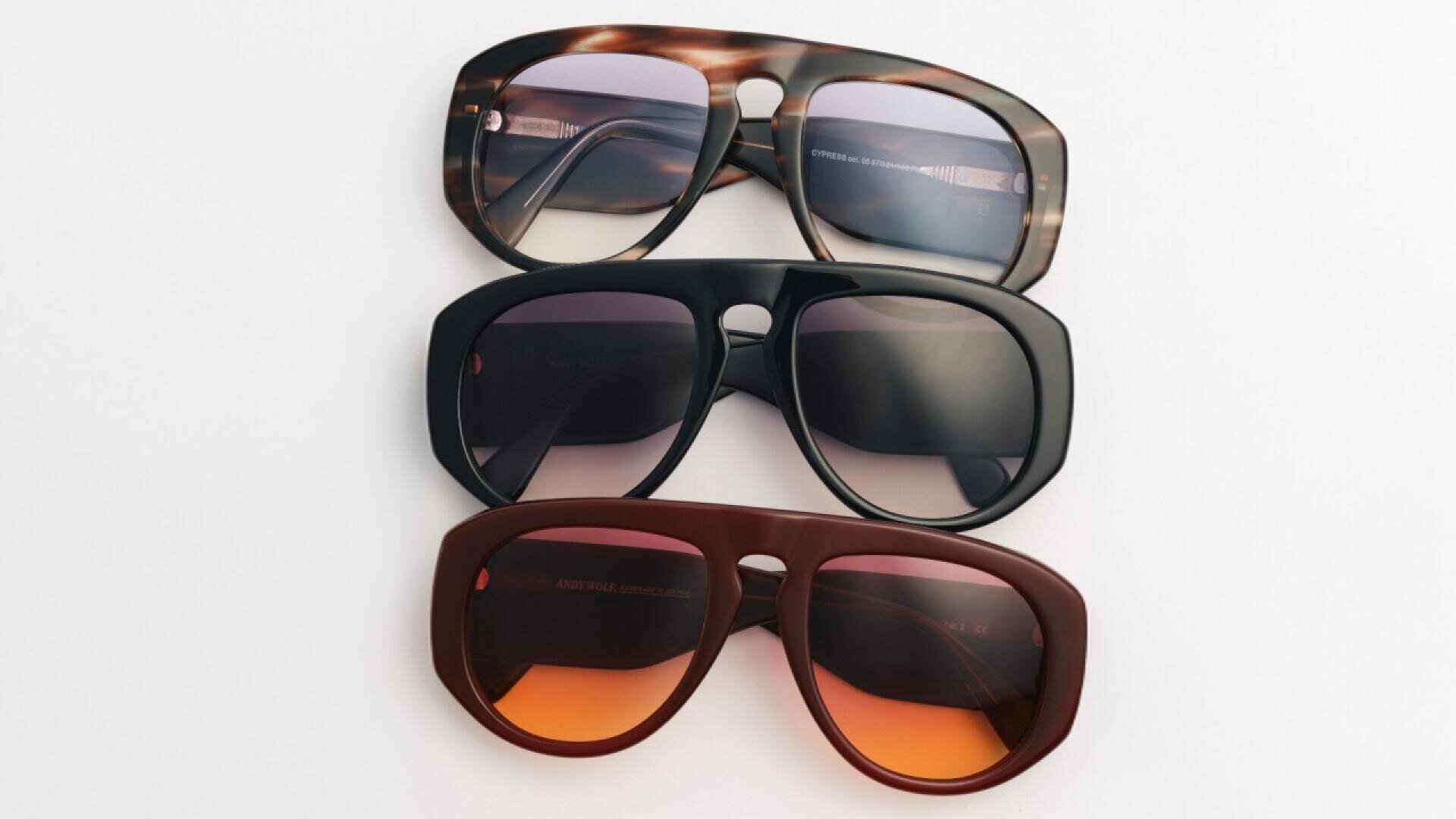 Three pairs of sunglasses with thick frames are stacked on a white surface. The top pair has a tortoiseshell pattern, the middle pair is black, and the bottom pair is dark red with orange-tinted lenses.