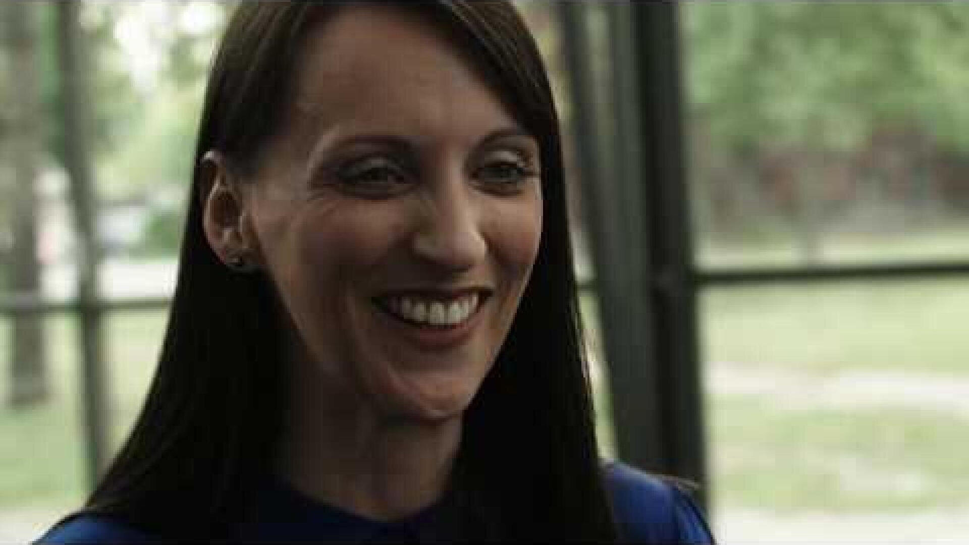 Eine Frau mit langen dunklen Haaren lächelt warm in einem Innenraum, in dem große Fenster und Grünflächen im unscharfen Hintergrund zu sehen sind.