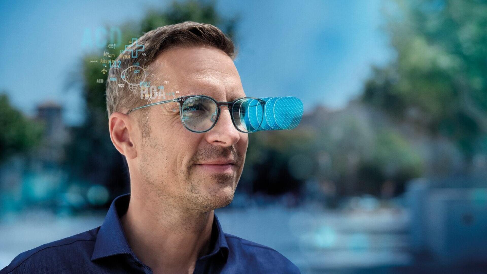 A man wearing glasses with futuristic digital graphics projected in front of his eyes, suggesting augmented reality technology, stands outdoors with a blurred background of trees and buildings.