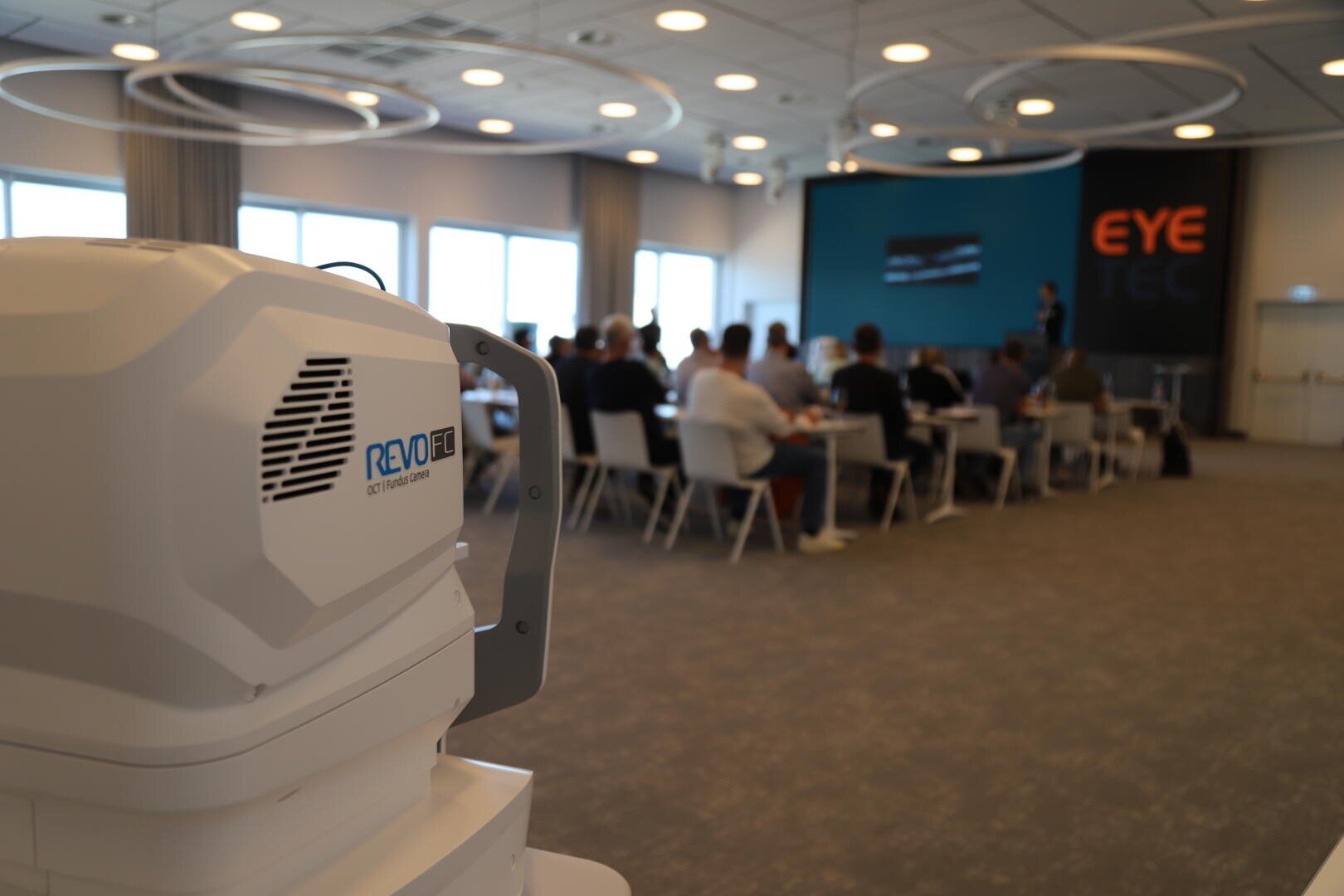 Im Vordergrund ist ein weißes medizinisches Gerät mit der Aufschrift REVO FC zu sehen, während eine Gruppe von Personen in einem modernen Konferenzraum mit der Aufschrift EYE TEC an der Rückwand vor einem Moderator und einem Bildschirm sitzt.