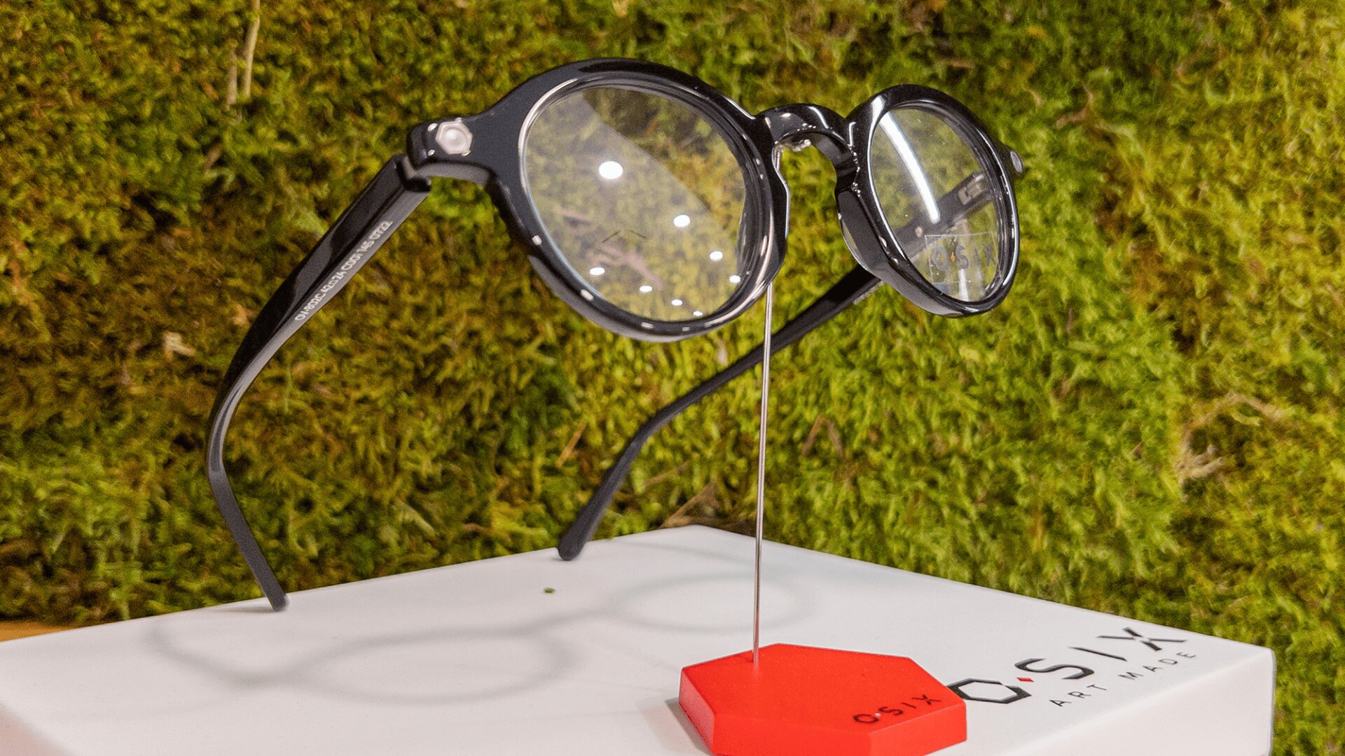 A pair of black round eyeglasses is displayed on a white stand with a red hexagonal base in front of a green mossy background. The word OSIX is partially visible on the stand.