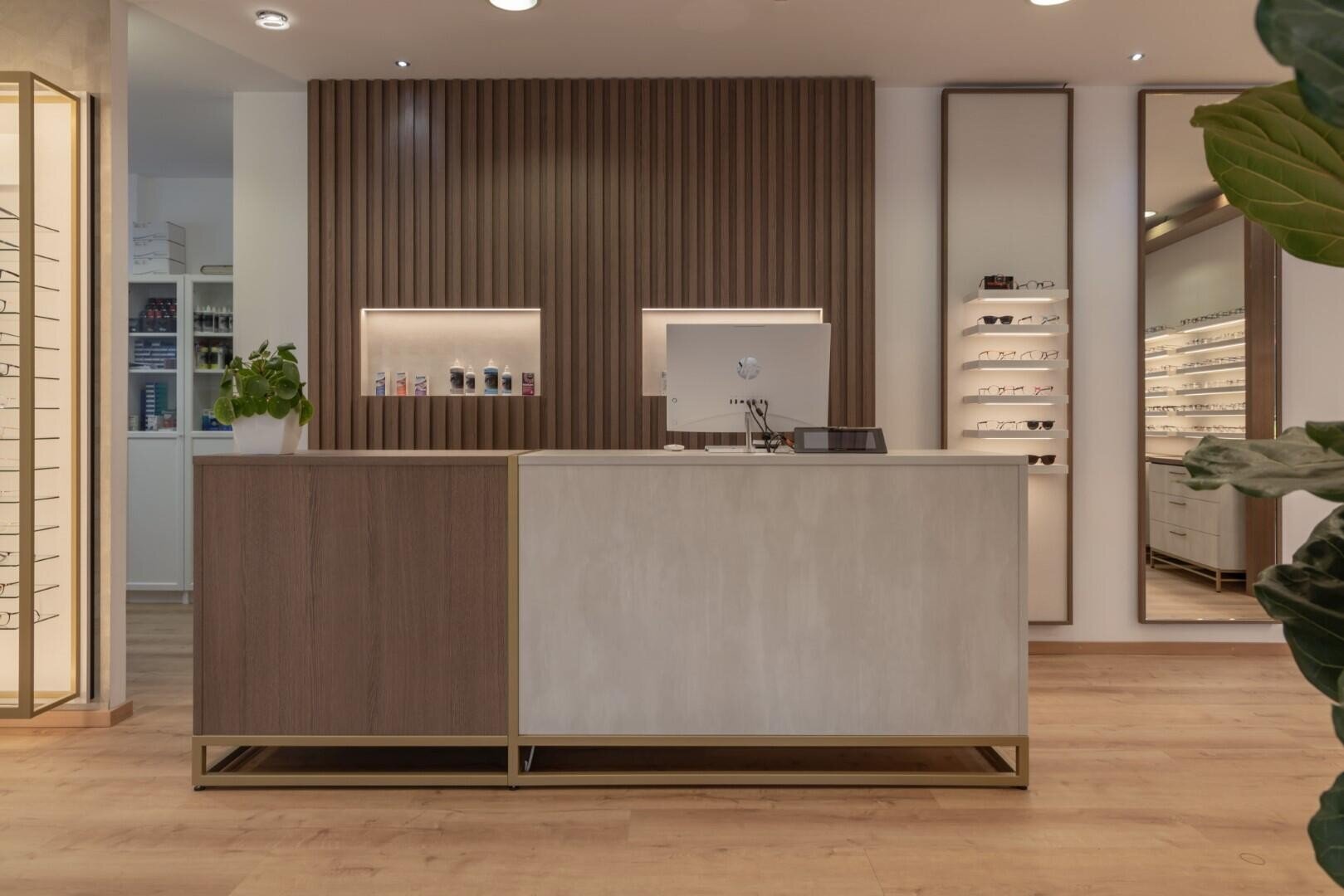 Ein moderner Empfangstresen eines Optikers mit Computer, Pflanzen und Regalen mit Brillen in einem gut beleuchteten Geschäft mit Holzböden und minimalistischem Dekor.