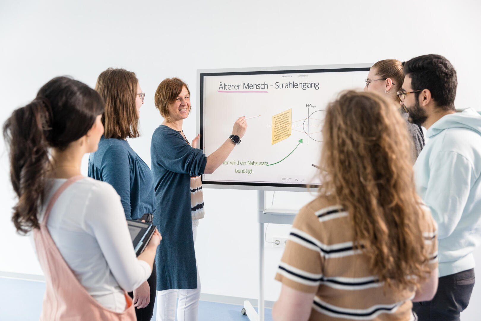 Eine Frau steht an einem digitalen Whiteboard und erklärt fünf aufmerksamen Erwachsenen in einem hellen Klassenzimmer ein Diagramm. Eine Person hält ein Tablet in der Hand, auf dem Bildschirm ist ein deutscher Text zum Thema „Älterer Mensch – Strahlengang“ zu sehen.