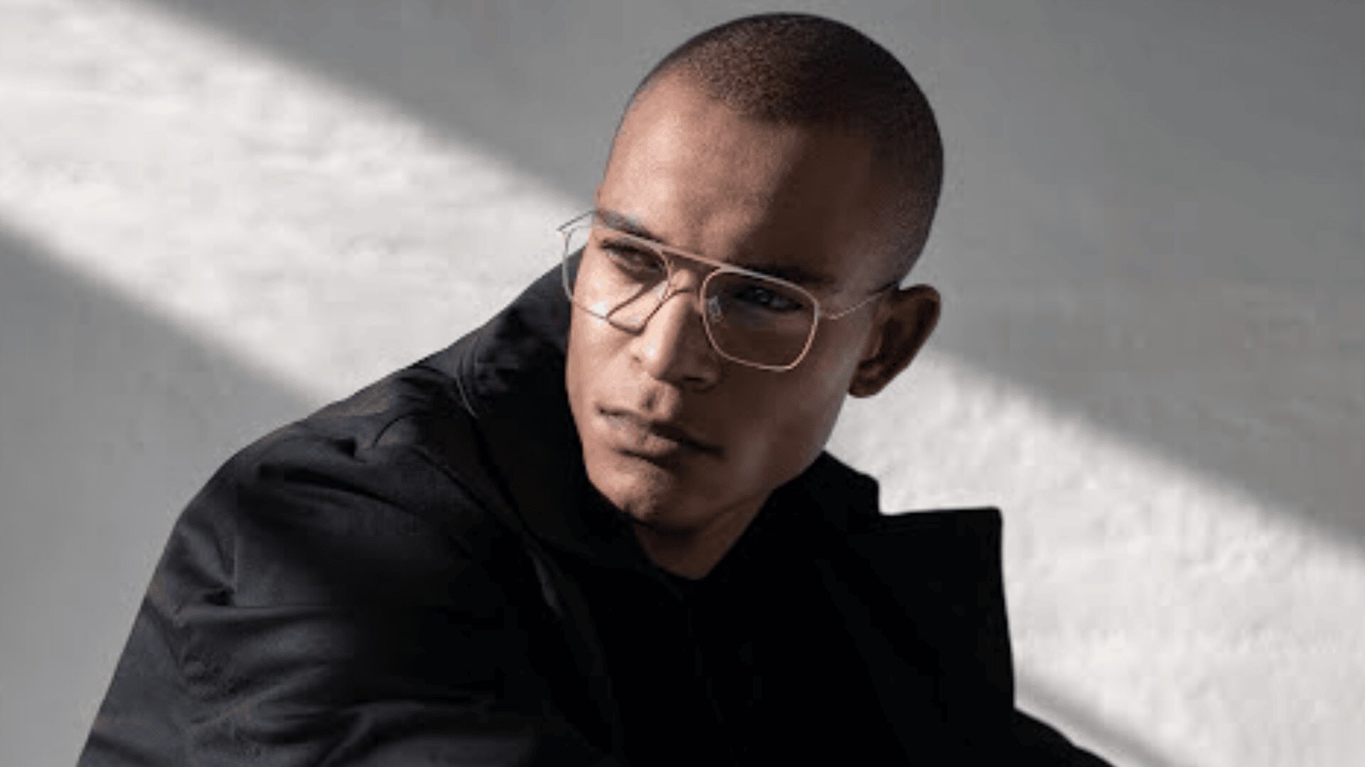 A man wearing clear-framed glasses and a dark jacket looks to the side in soft, natural light, with shadows cast on the wall behind him.