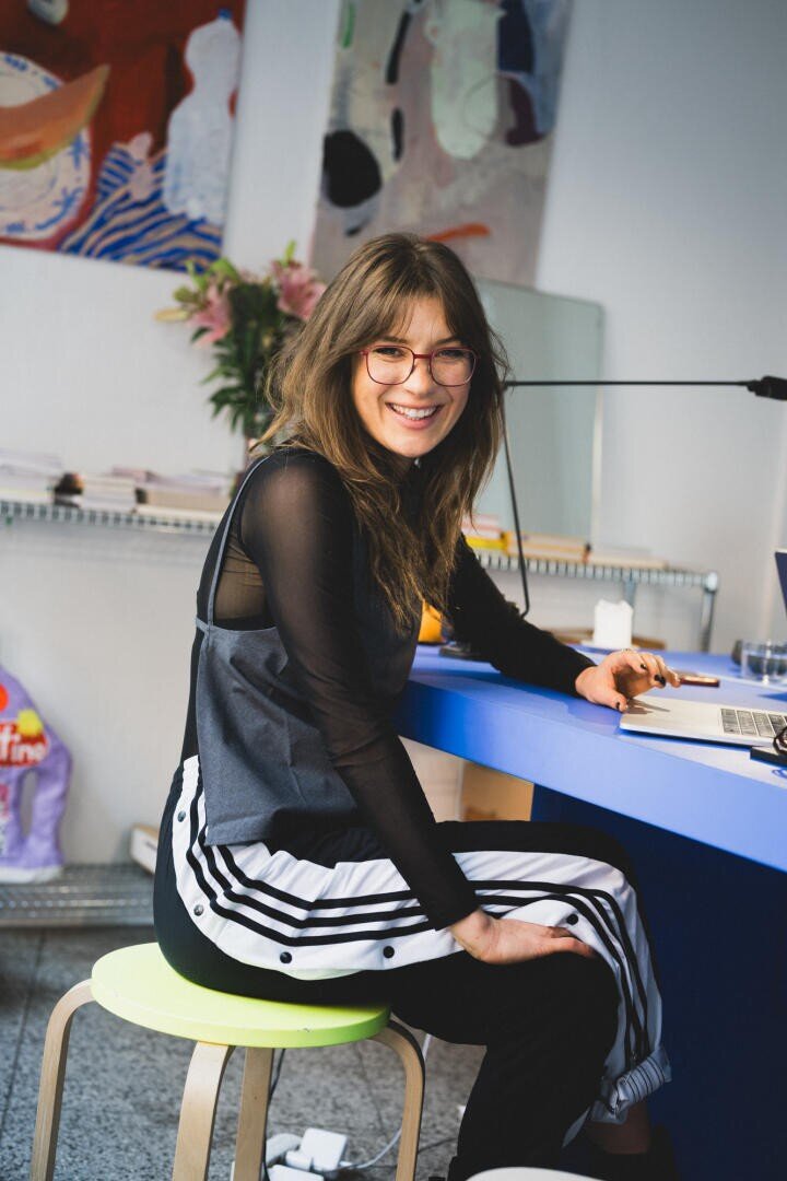 Eine Frau mit langen braunen Haaren und Brille sitzt lächelnd an einem blauen Schreibtisch in einem modernen, hell erleuchteten Büro und trägt ein schwarzes durchsichtiges Oberteil und eine sportlich gestreifte Hose. Im Hintergrund sind bunte Kunst und Blumen zu sehen.