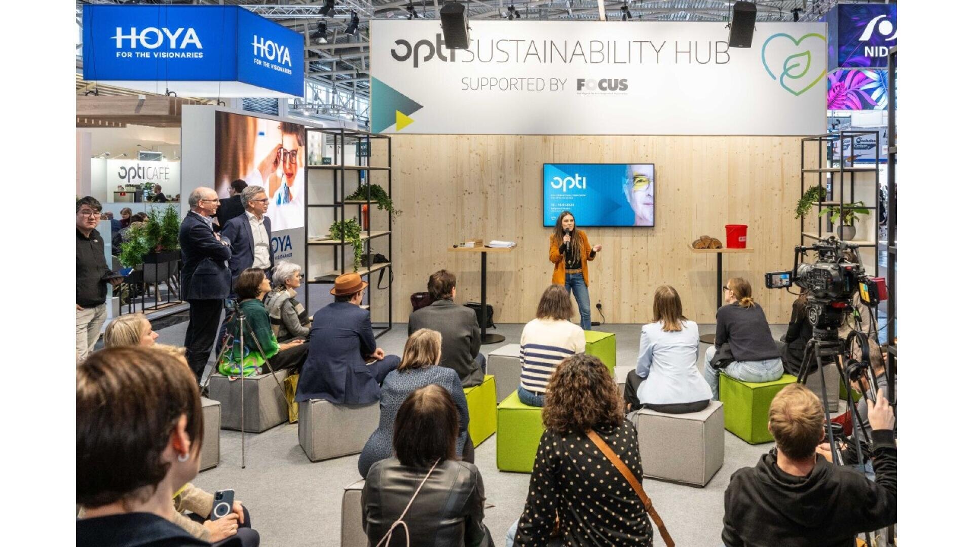 A woman speaks on stage at the OPTI Sustainability Hub, attended by a seated audience. The event is indoors, with banners, cameras, and a wooden backdrop displaying event sponsors.