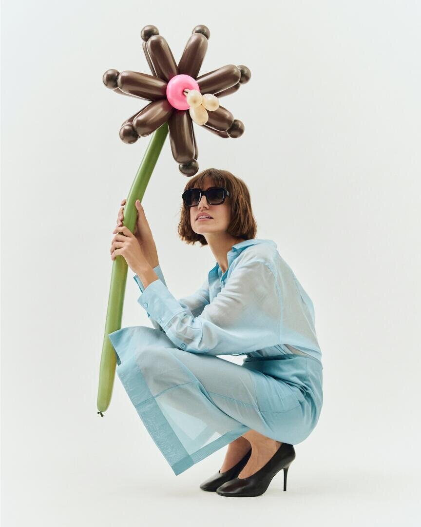 A woman with sunglasses, a light blue see-through dress and black heels squats and holds a large balloon flower with dark petals and a green stem in front of a plain white background.
