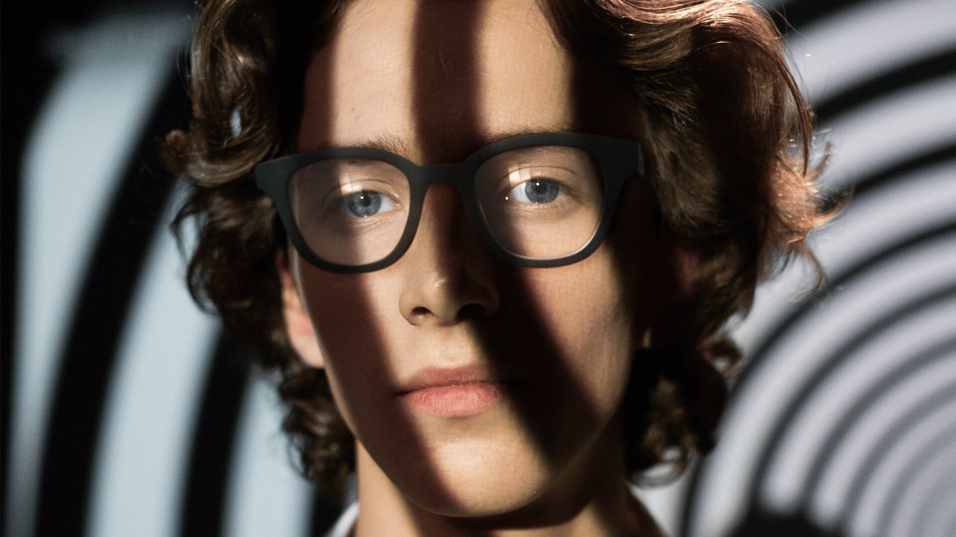 Eine Person mit lockigem Haar und Brille steht vor einem schwarz-weißen Spiralhintergrund, wobei Schatten dunkle Streifen über ihr Gesicht werfen.