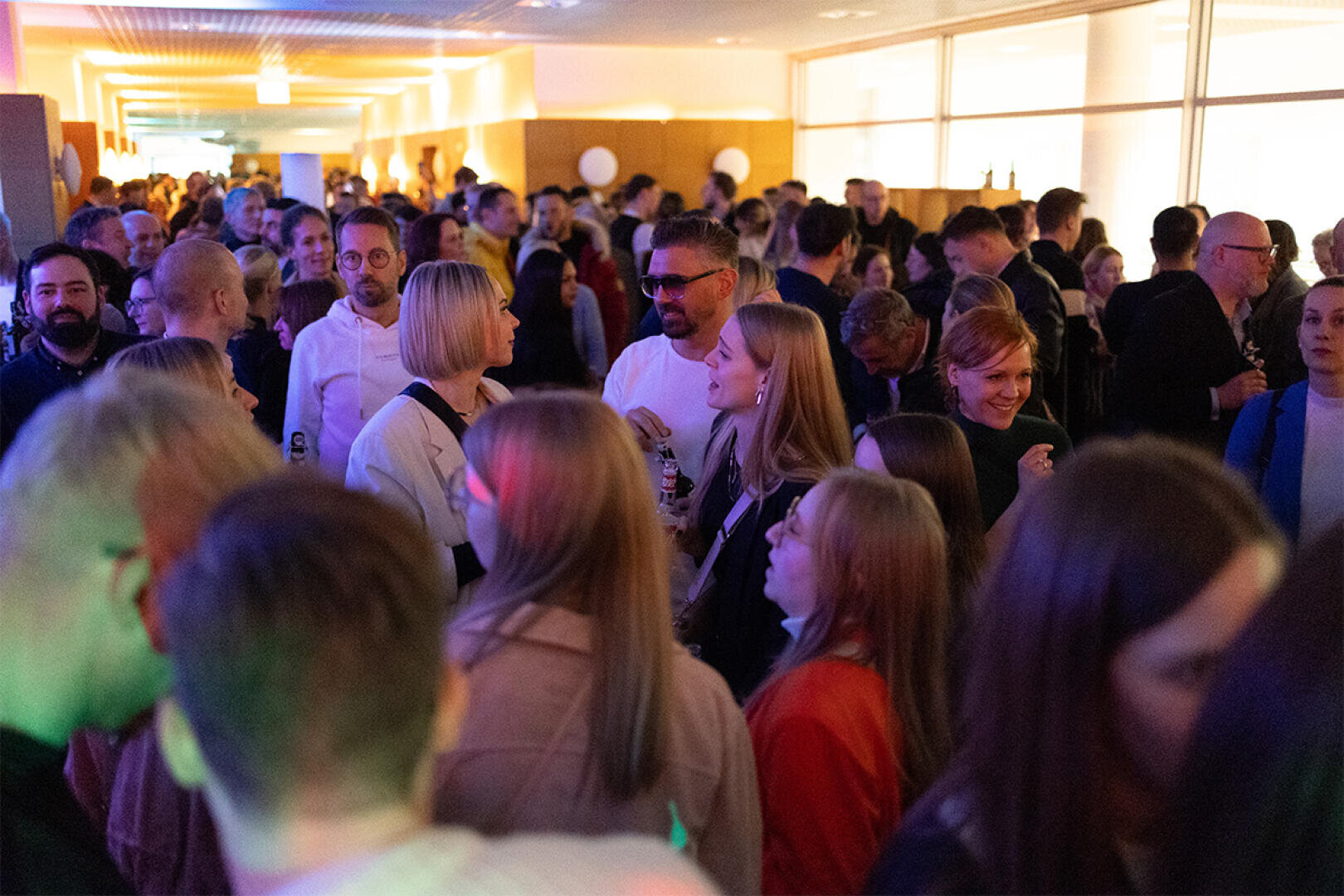 Eine große Gruppe von Menschen trifft sich während einer Veranstaltung in einem hell erleuchteten Flur, viele unterhalten sich und einige halten Getränke in der Hand. Die Atmosphäre wirkt lebendig und überfüllt.