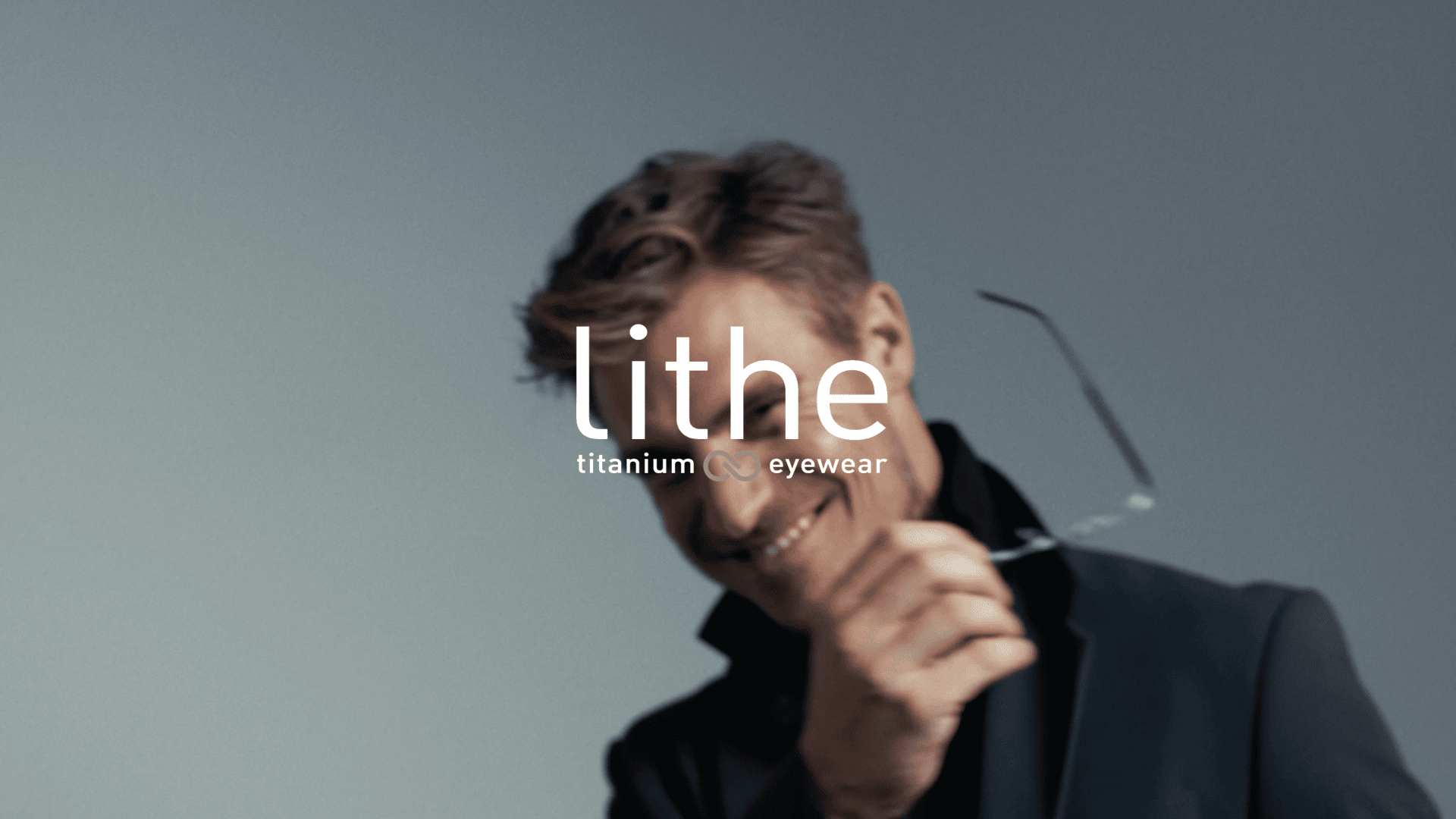 A man in a dark blazer smiles while removing eyeglasses, with the words “lithe titanium eyewear” displayed over the image. The background is plain and softly lit.