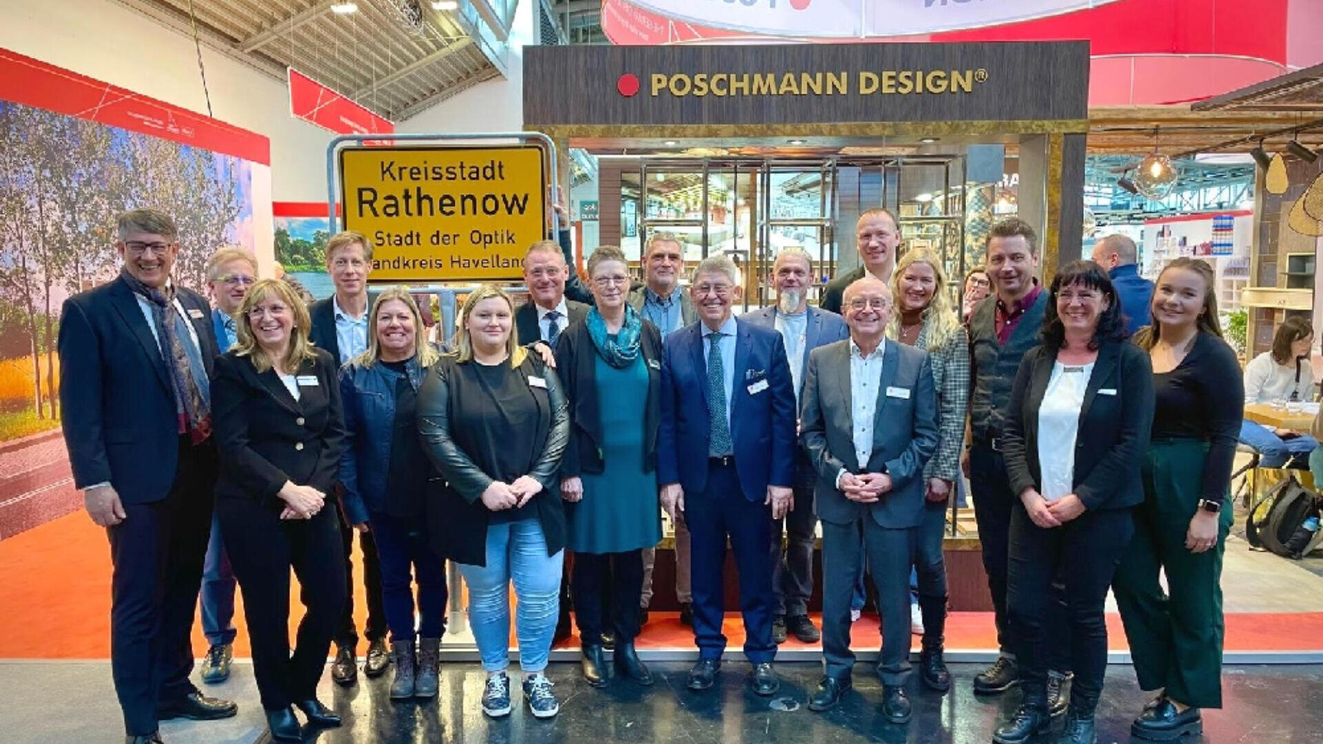 A group of people stand smiling in front of a booth with a “Kreisstadt Rathenow” sign and a Poschmann Design banner, at what appears to be an indoor exhibition or trade fair.