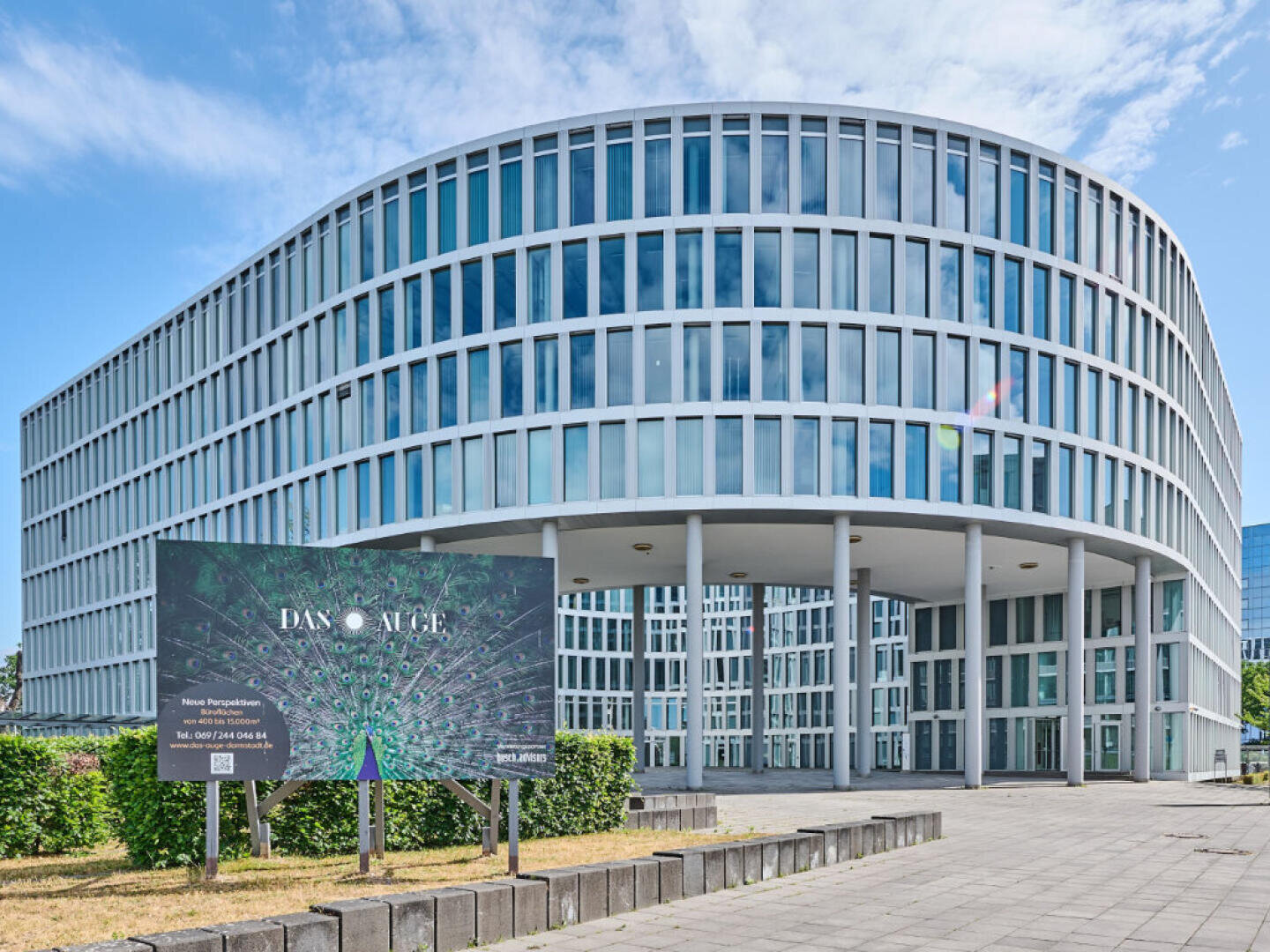 A modern, curved office building with many windows stands behind a sign displaying “DAX Allee” and an abstract graphic. The sky is partly cloudy and the area is paved with some greenery in front.