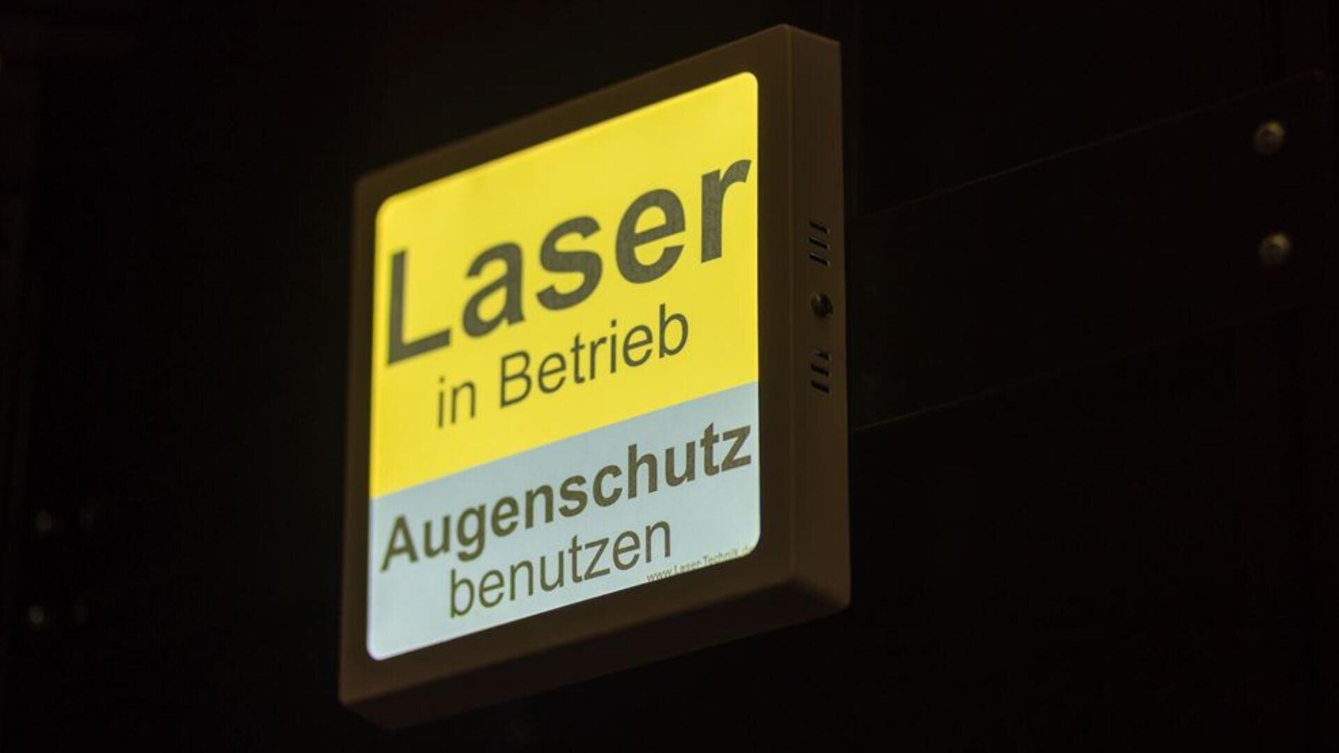 Auf einem gelb-weißen Warnschild steht in deutscher Sprache „Laser in Betrieb, Augenschutz benutzen“, was bedeutet, dass der Laser in Betrieb ist und ein Augenschutz verwendet werden muss. Das Schild leuchtet vor einem dunklen Hintergrund.