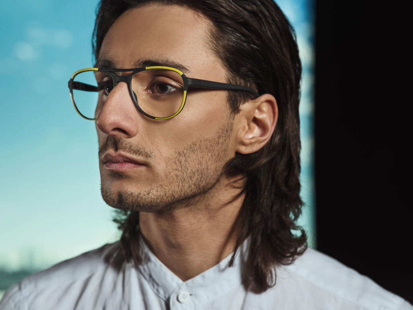 Ein junger Mann mit langen dunklen Haaren und einer gelb umrandeten schwarzen Brille blickt zur Seite. Er trägt ein weißes Hemd und steht vor einem sanften blauen Hintergrund mit einer verschwommenen Stadtlandschaft.