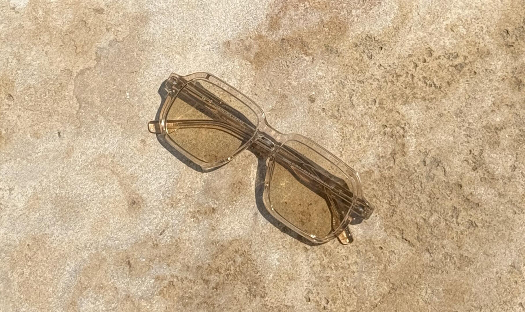 Transparent brown sunglasses with a rectangular frame lie under bright sunlight on a textured, light brown stone surface.