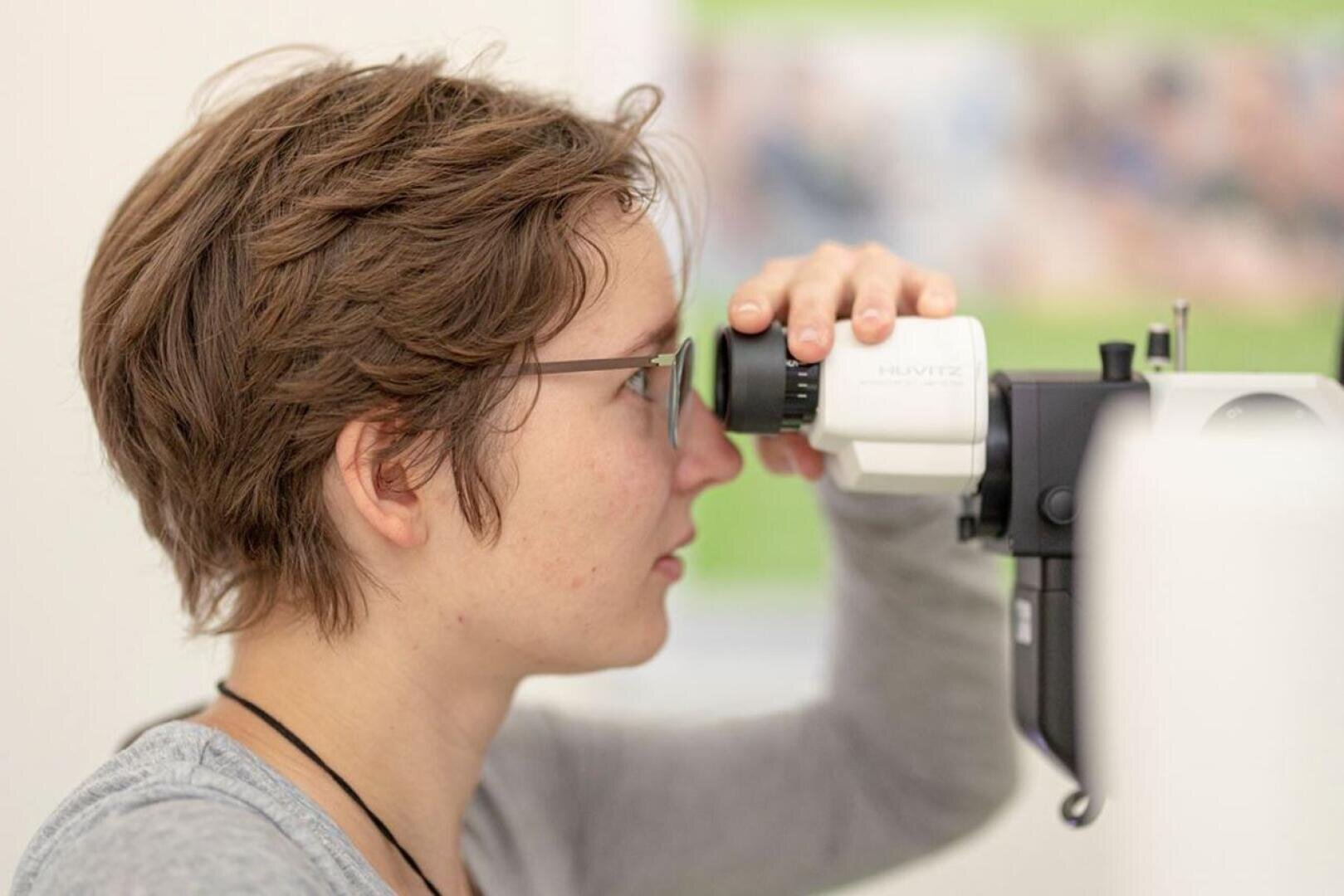 Eine Person mit kurzen braunen Haaren und Brille sitzt im Profil und blickt während eines Sehtests in einem gut beleuchteten Raum in ein Augenuntersuchungsgerät.