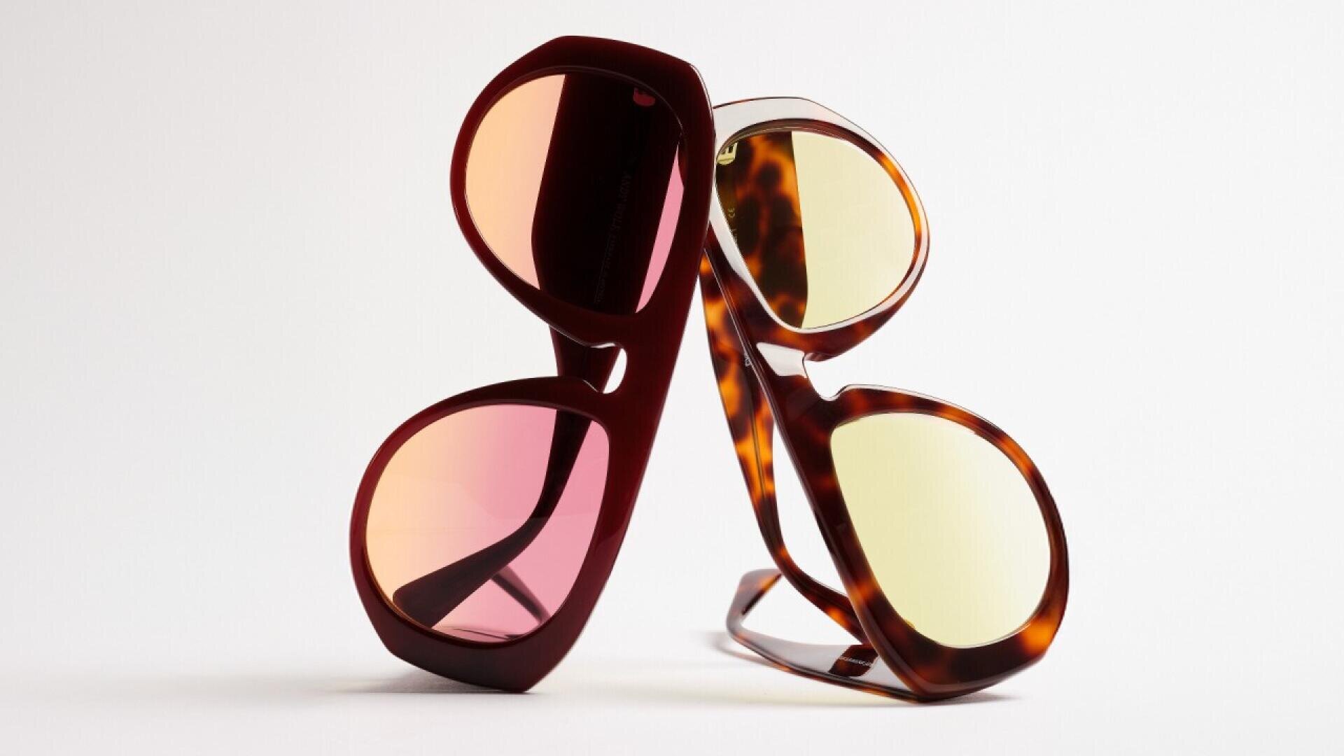 Two pairs of stylish sunglasses with oversized frames, one with pink-tinted lenses and maroon frames, the other with yellow-tinted lenses and tortoiseshell frames, are leaning against each other on a white background.
