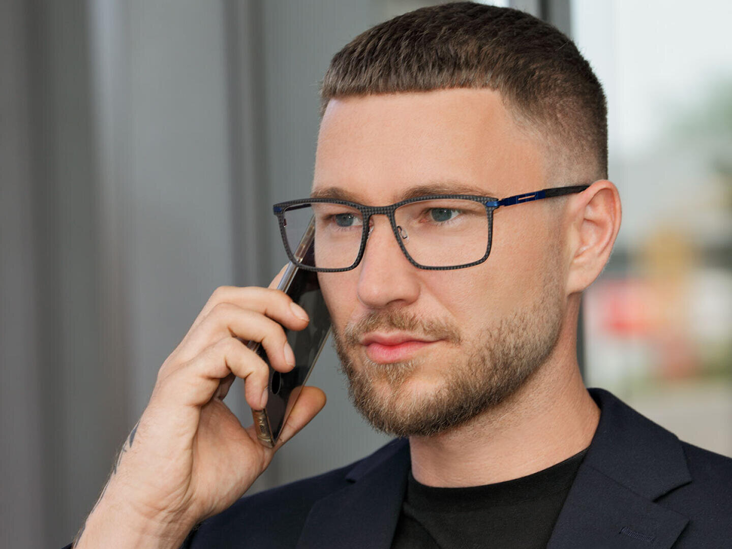 Ein Mann mit kurzen Haaren und Bart, Brille und dunklem Blazer hält ein Smartphone ans Ohr und scheint zu telefonieren.