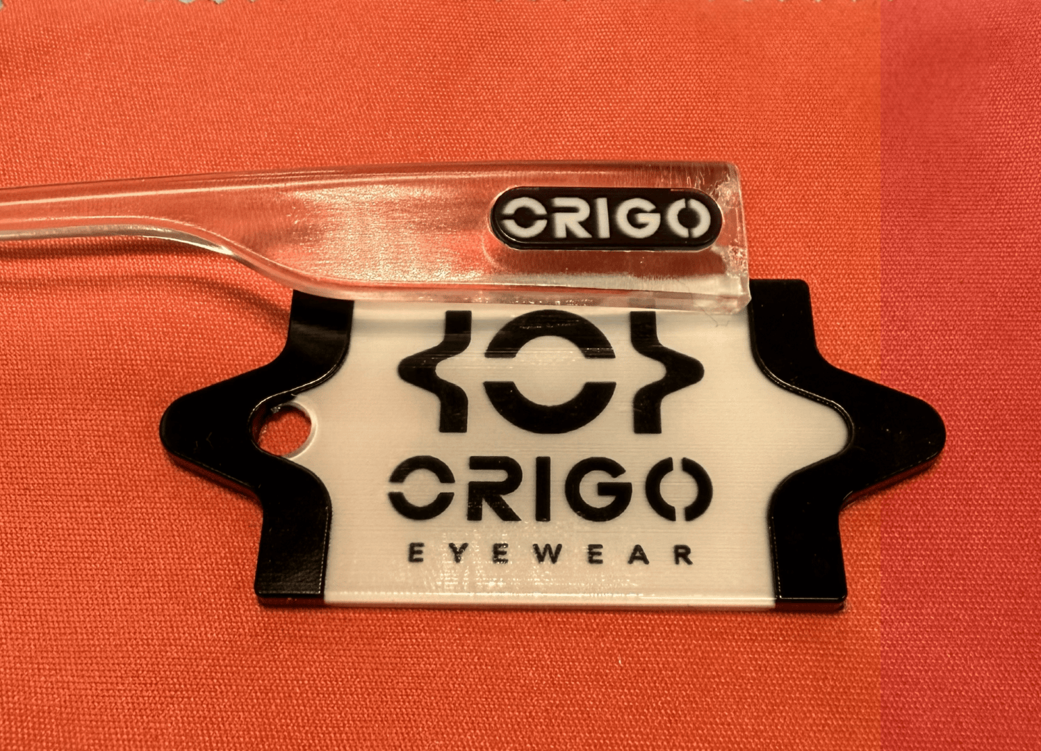 Ein durchsichtiger Brillenbügel mit dem ORIGO-Logo ruht auf einer weiß-schwarzen Displaykarte der Marke ORIGO EYEWEAR, die auf einer rot-orangen Stoffoberfläche platziert ist.