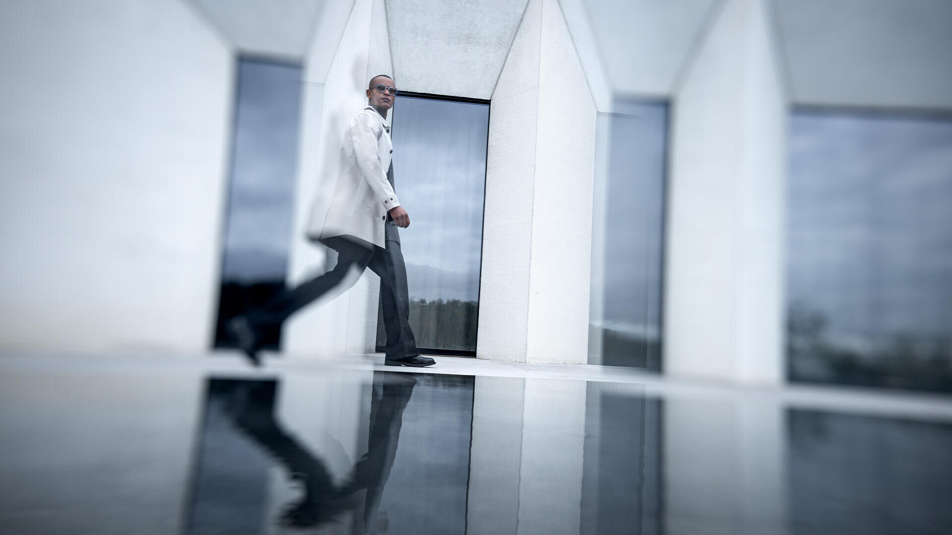 Ein Mann in einem hellen Mantel und einer dunklen Hose geht zügig vor einem modernen weißen Gebäude mit hohen Fenstern, sein Spiegelbild ist in einer glänzenden Oberfläche darunter zu sehen. Das Bild ist leicht unscharf, um die Bewegung zu betonen.