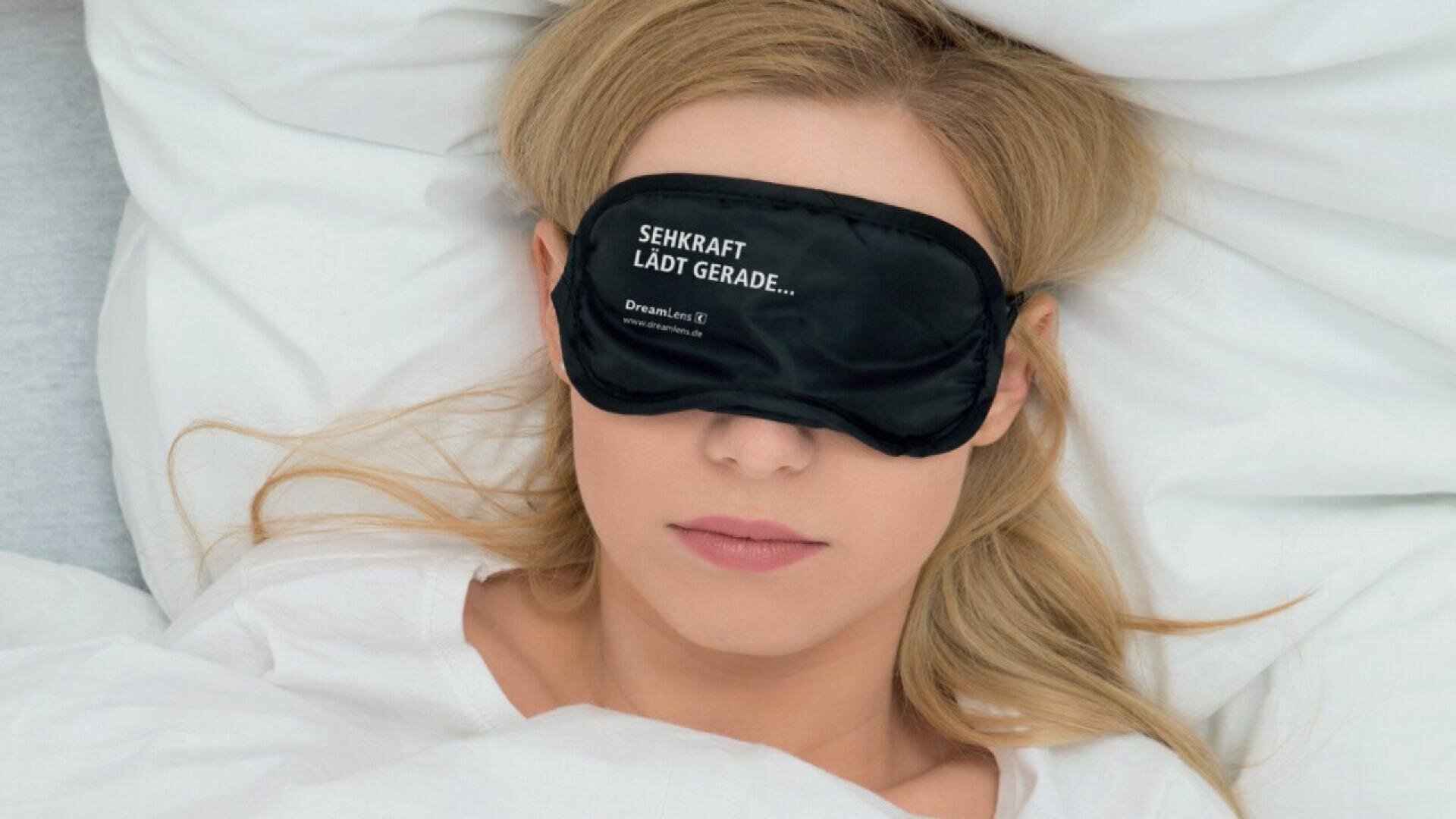 A woman lies in bed with white sheets, wearing a black sleep mask that has German text on it. Her eyes are covered and she appears to be resting or sleeping.