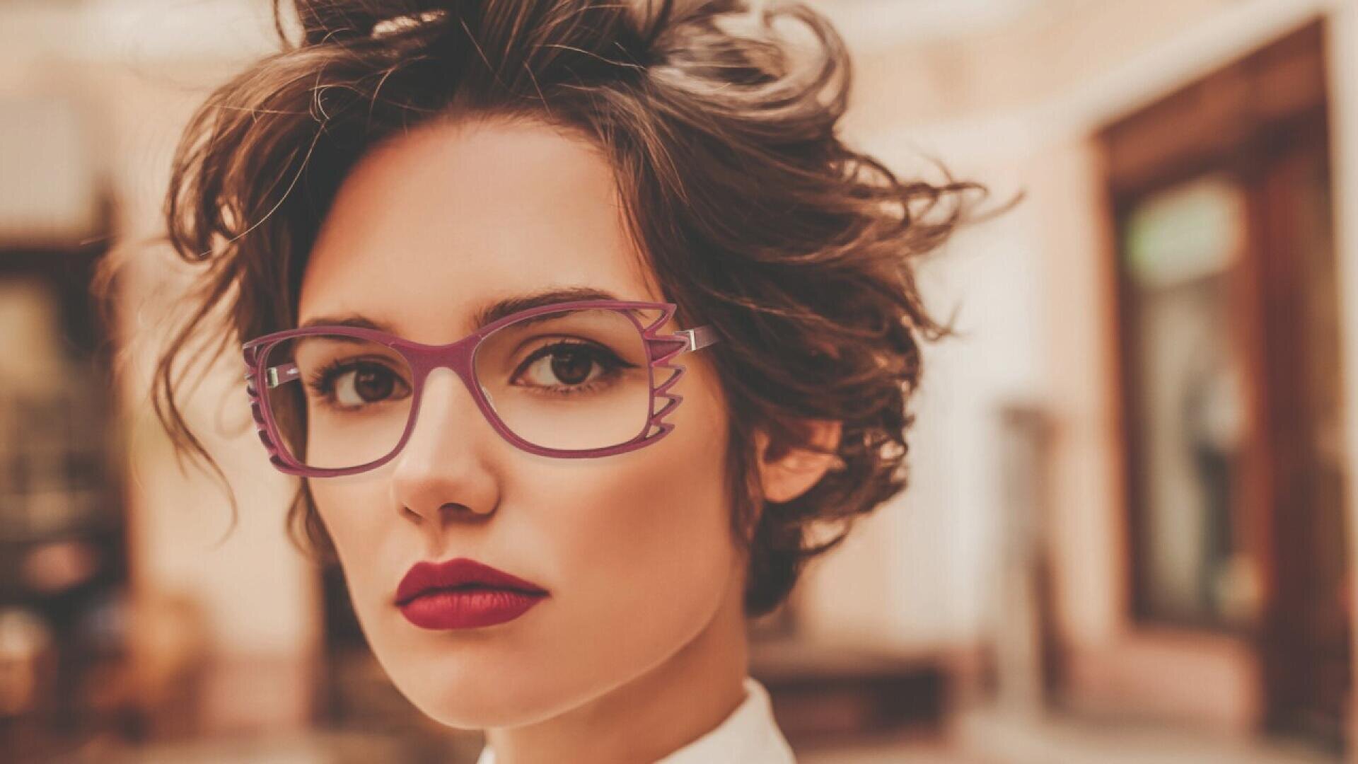 A woman with short, wavy brown hair wears bold, purple glasses with decorative frames and red lipstick, looking confidently at the camera. The background is softly blurred.