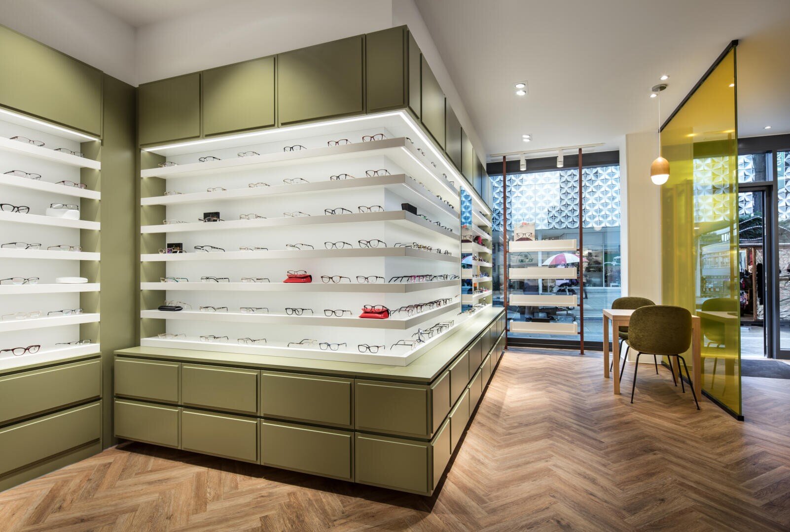 Ein modernes Brillengeschäft mit olivgrünen Regalen, in denen verschiedene Brillen ausgestellt sind. Der Raum verfügt über einen Fischgrätenholzboden, eine Sitzecke mit Stühlen und große Fenster, die natürliches Licht hereinlassen.
