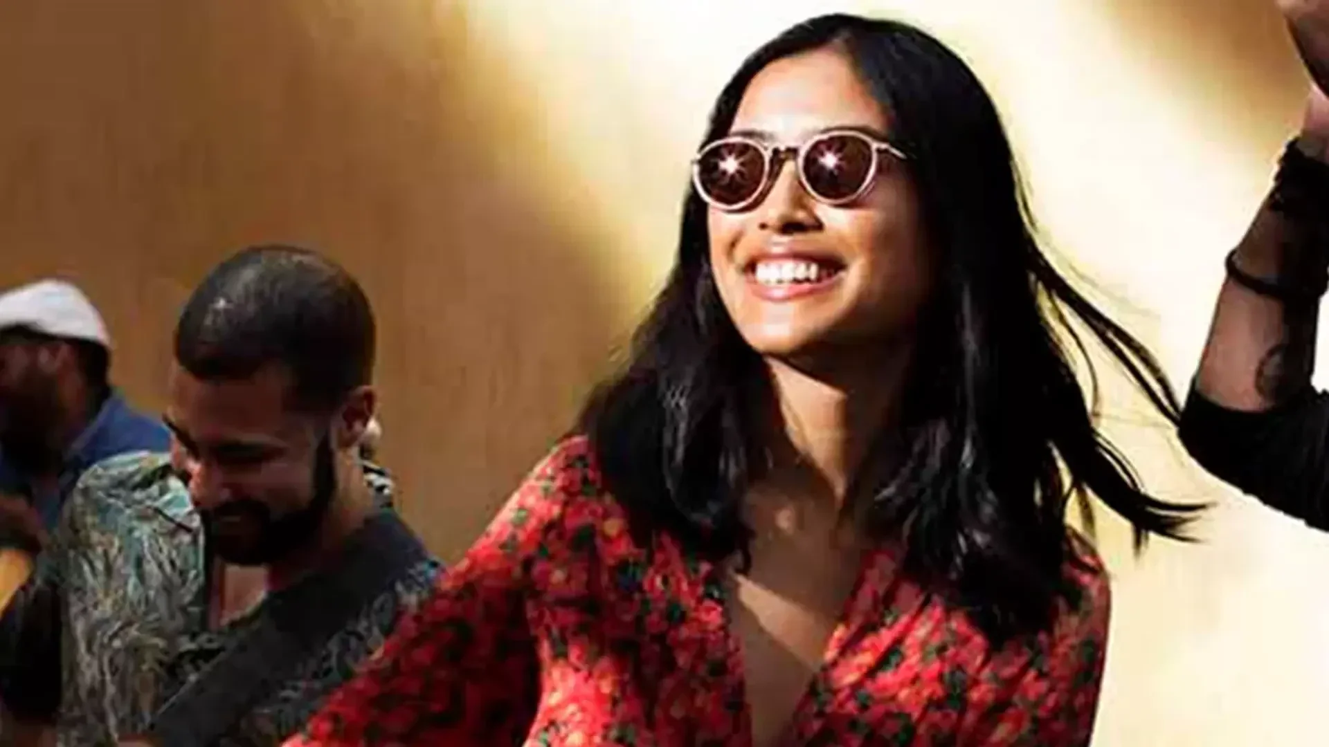 Eine Frau mit Sonnenbrille und rotem Blumentop lächelt strahlend im Sonnenlicht, im Hintergrund spielt ein Mann Gitarre.